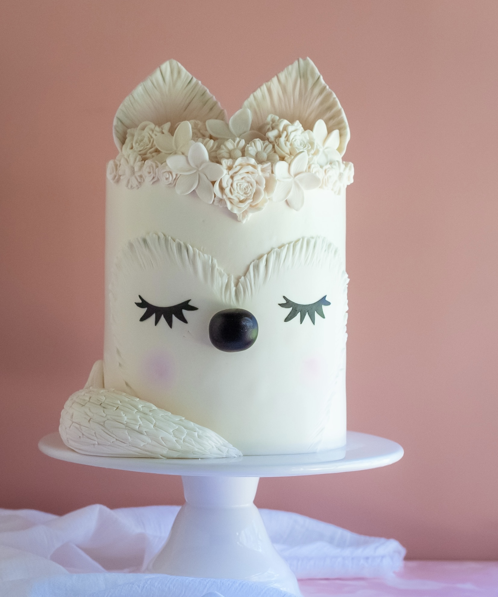 Cake decorated as a fox with closed eyes, black nose, floral ears, and a pink background.