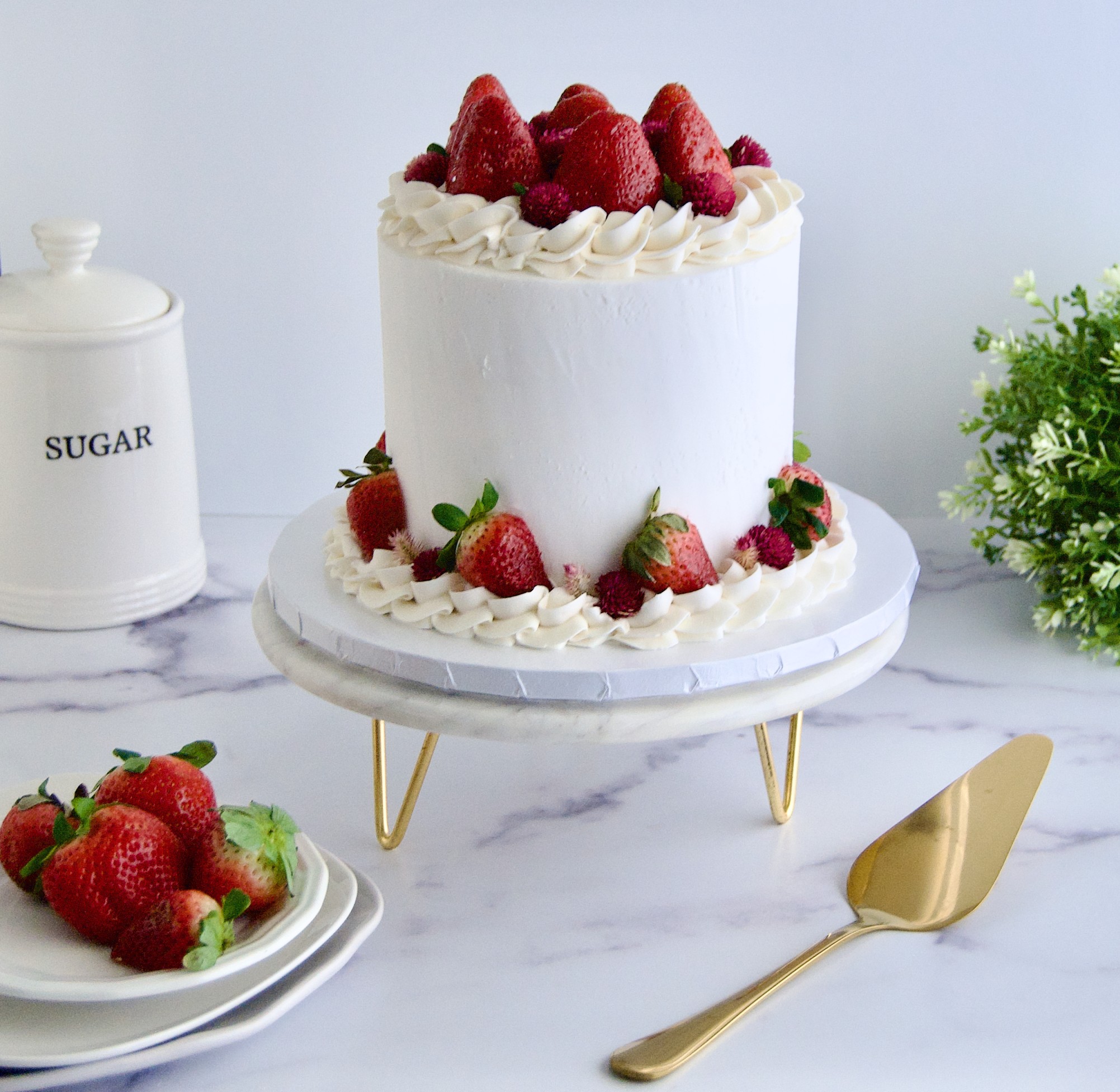 White cake with strawberries on top and around, on a white cake stand, with a plate of strawberries and a gold cake server nearby.