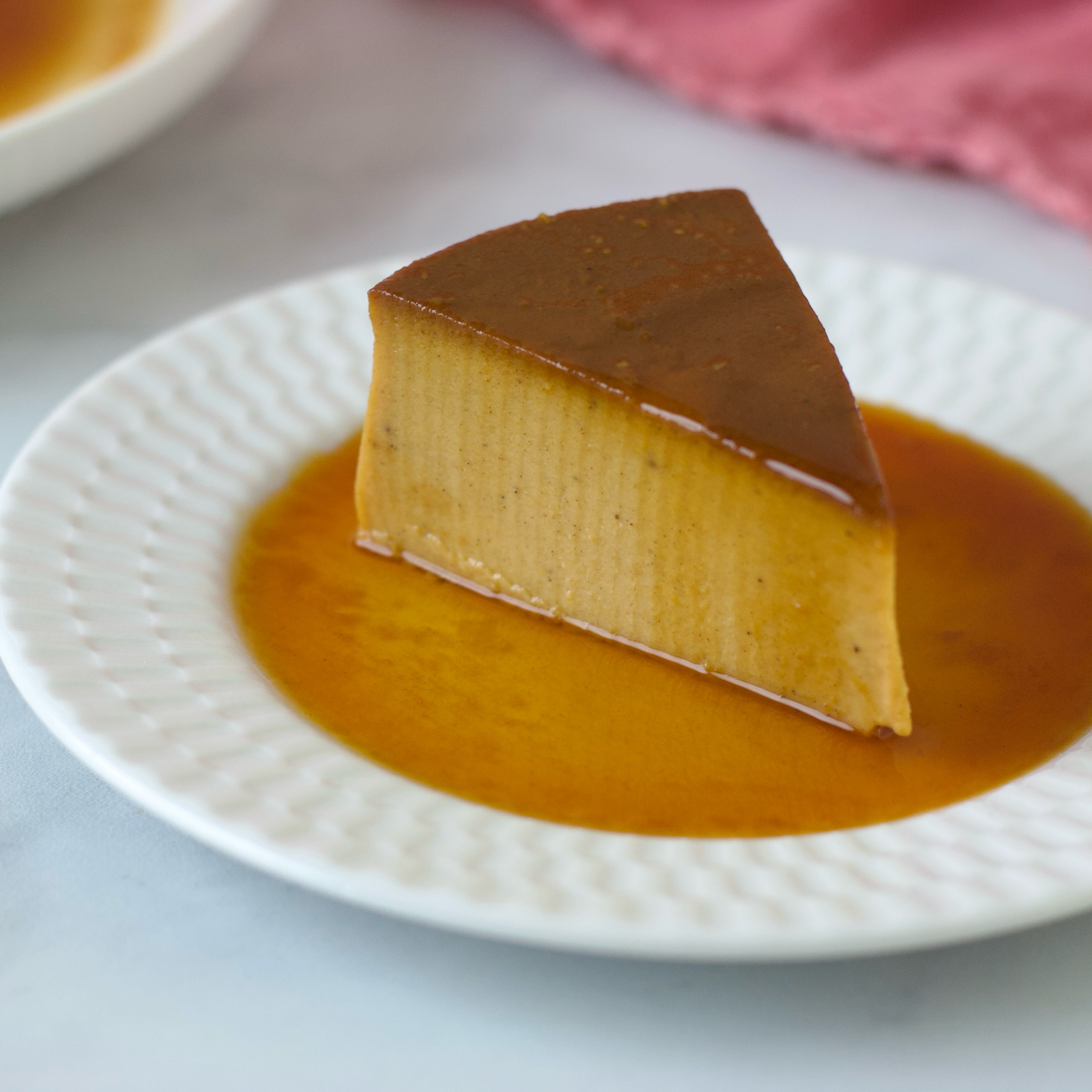 Slice of flan with caramel sauce on a white textured plate, with a pink cloth in the background.