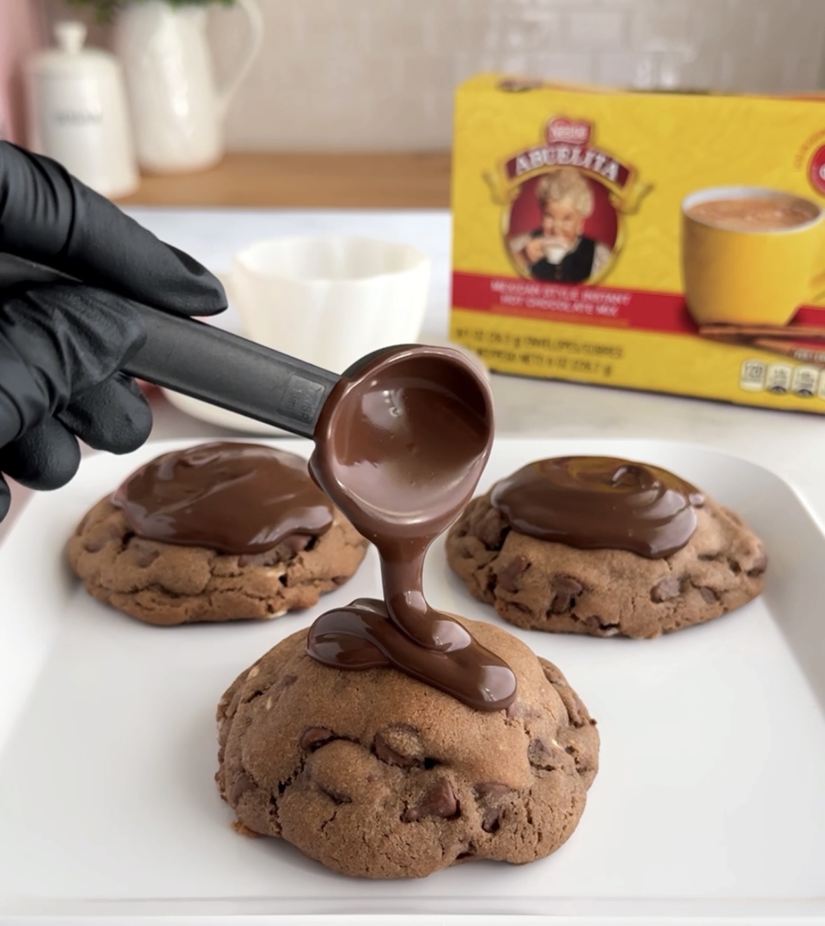 Hand in black glove drizzling melted chocolate onto cookies with chocolate topping, on a white plate, with cookie mix box in background.