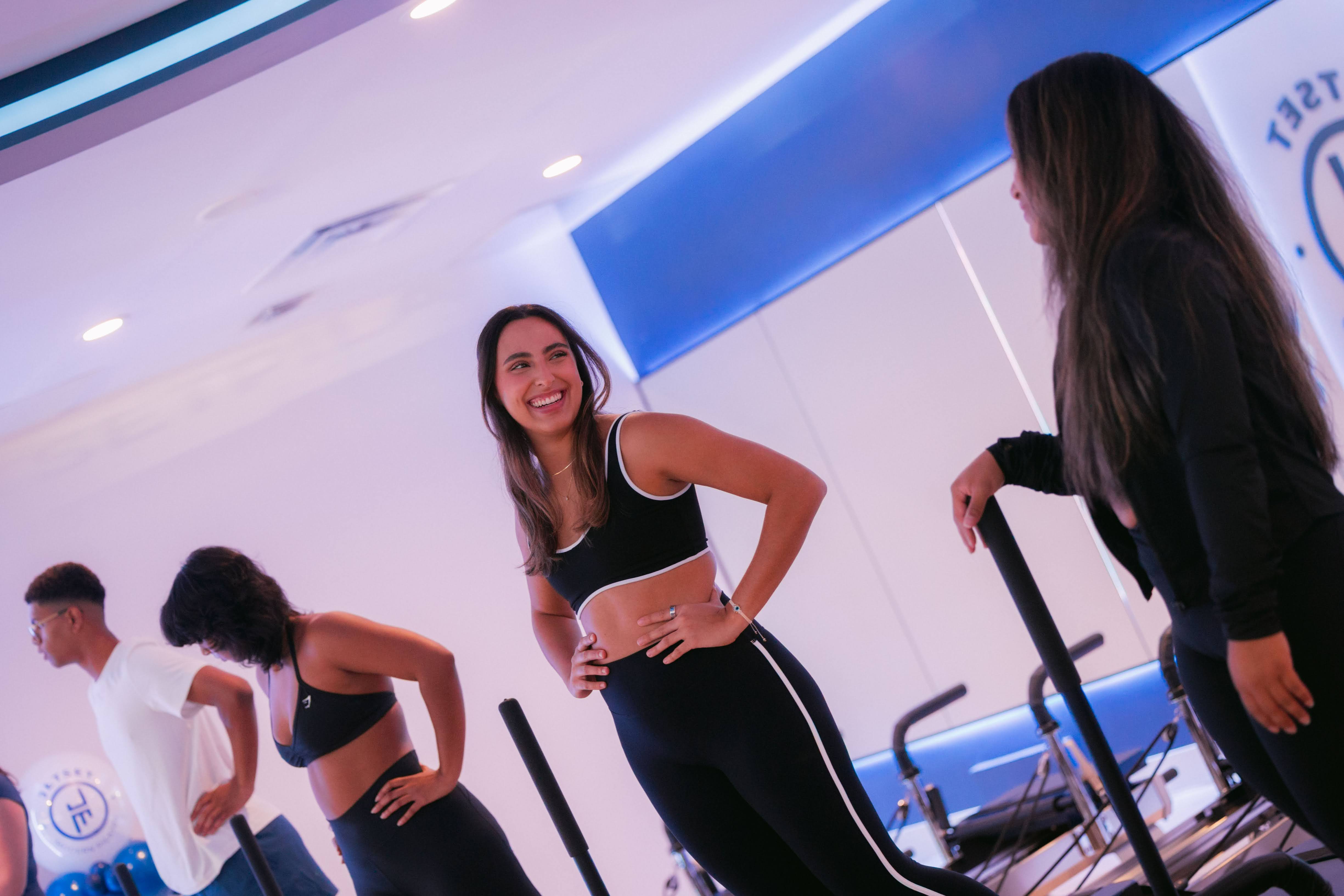 Two women and a man in athletic clothing standing and talking indoors, with exercise equipment in the background.