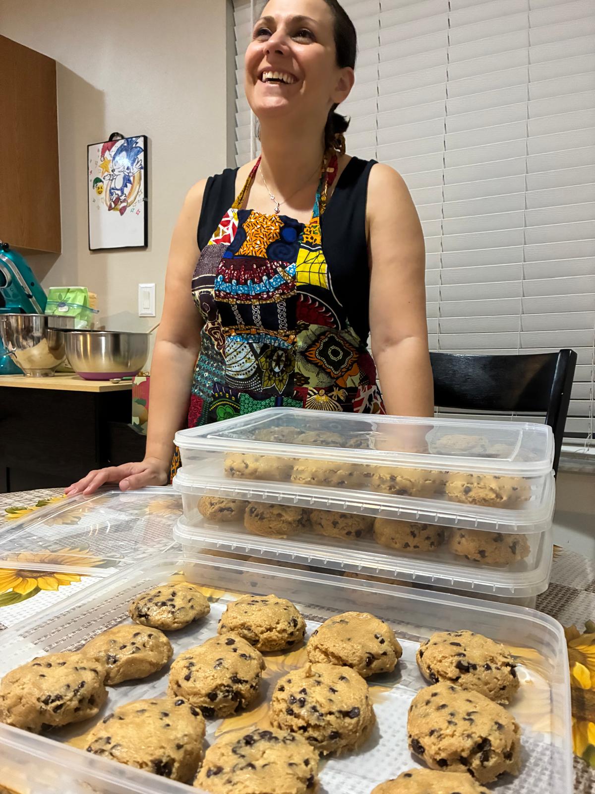 Smiling woman wearing an apron stands behind stacked containers of cookies in a kitchen.