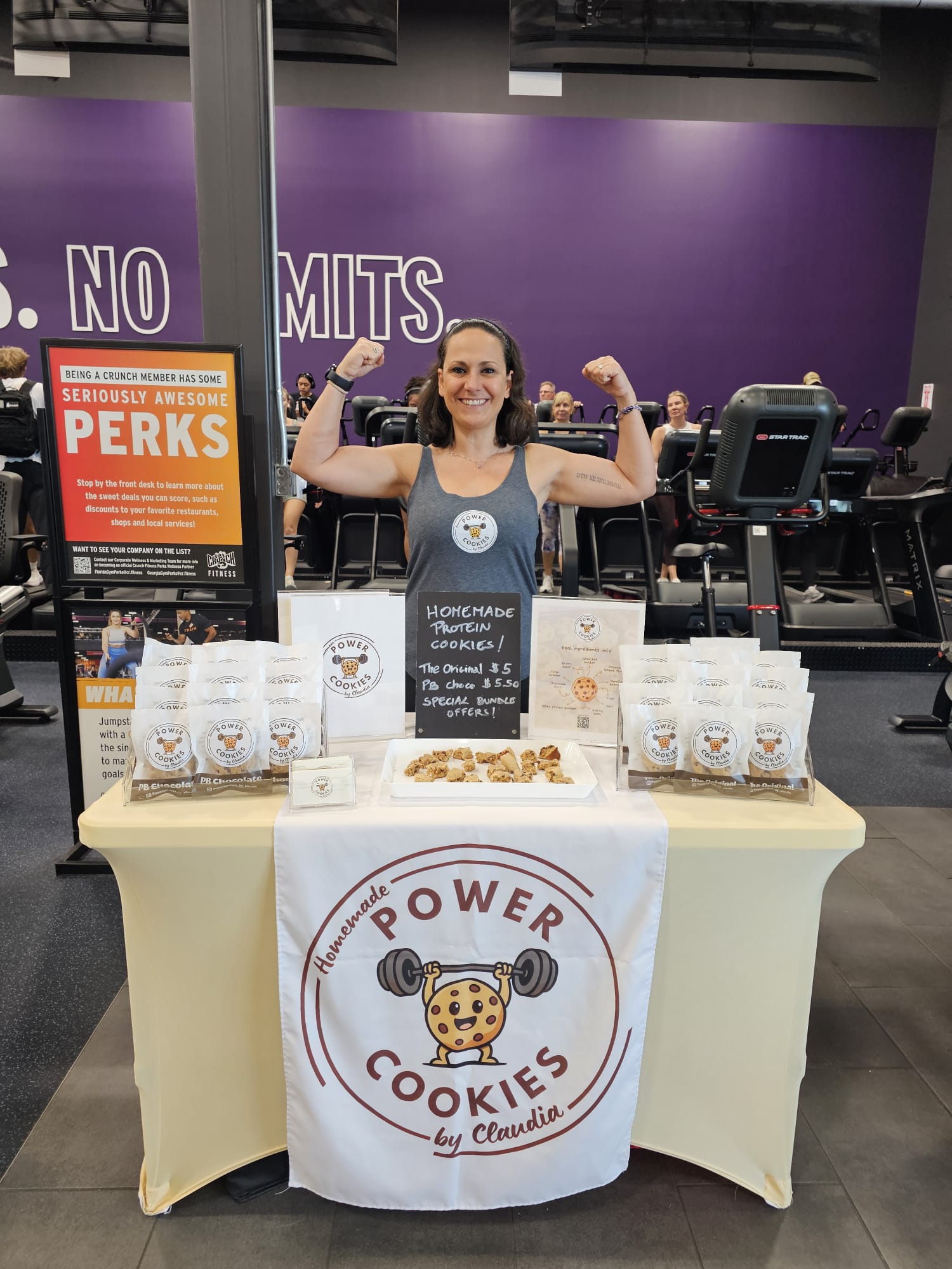 Woman standing behind a table with cookies and promotional materials in a gym, smiling and flexing arms, purple wall in background.