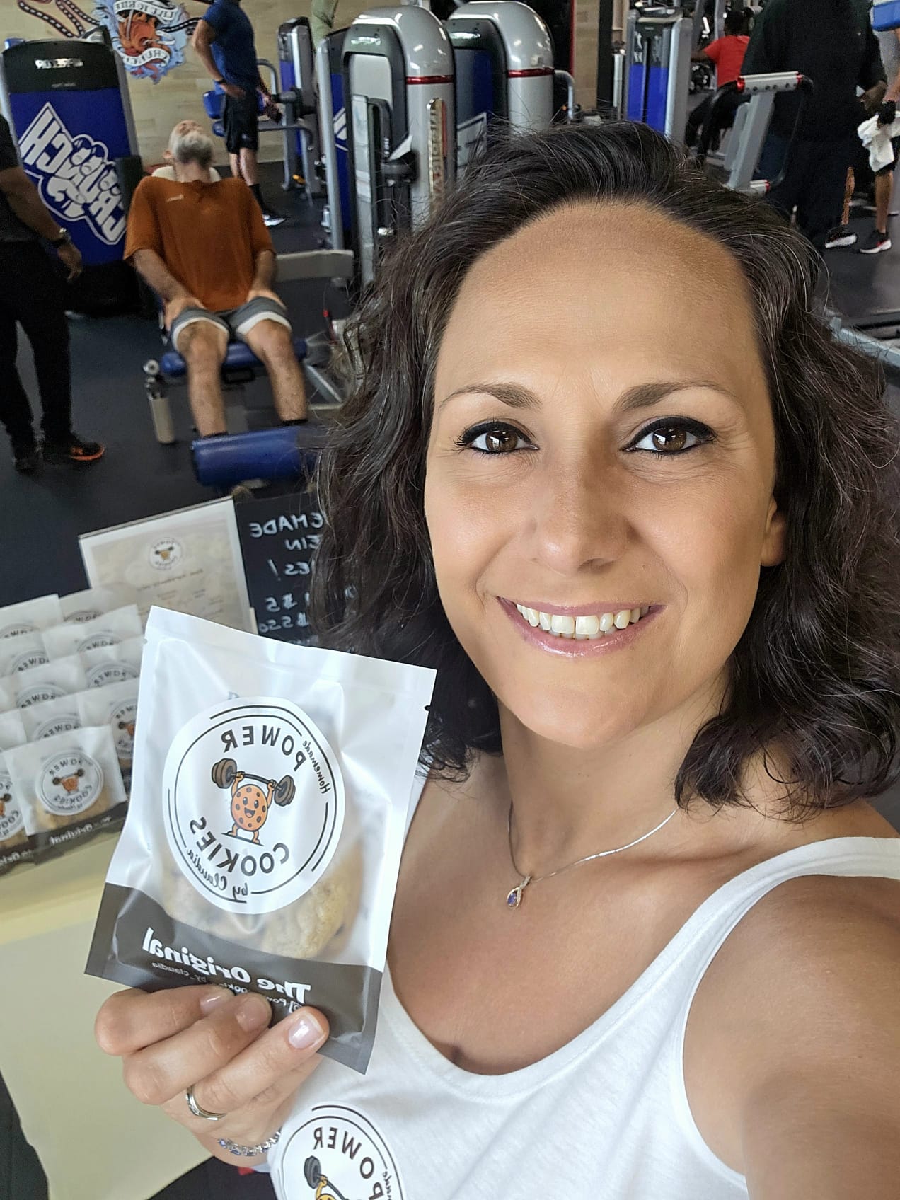 Woman smiling in gym holding packaged cookie, gym equipment in background.