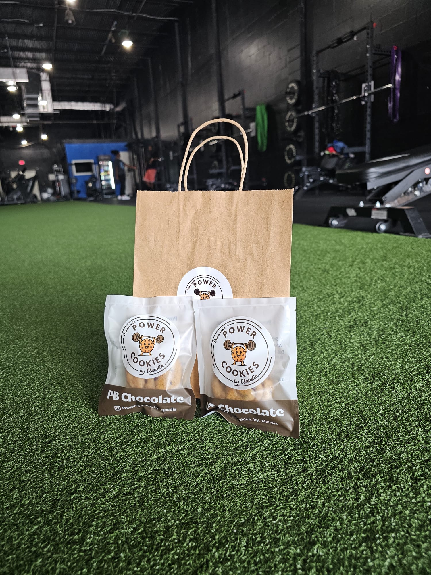 Two cookies in white packaging with a bear logo, a brown paper bag, and a black ribbon on green artificial turf inside a gym.
