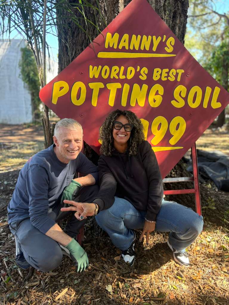 Two people crouching in front of a tree with a large red sign advertising potting soil, outdoors with trees and sunlight.