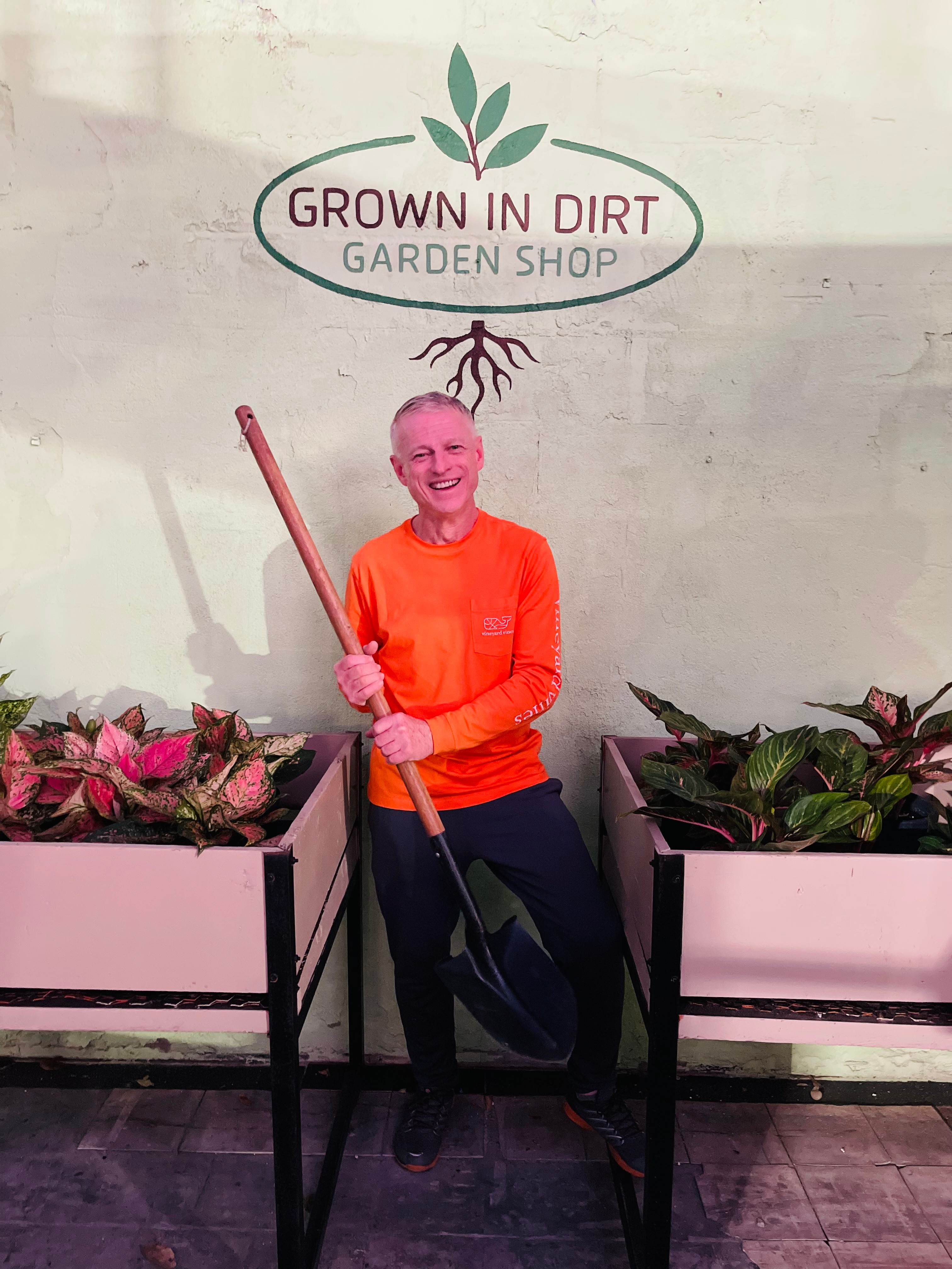 Person sitting between two pink planters with plants, holding a gardening tool, smiling, with a sign above reading 'GROWN IN DIRT GARDEN SHOP' and a root illustration.