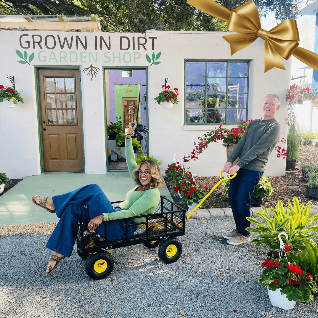 Two women with one pulling a cart in front of a garden shop, surrounded by flowers and greenery, sunny day.