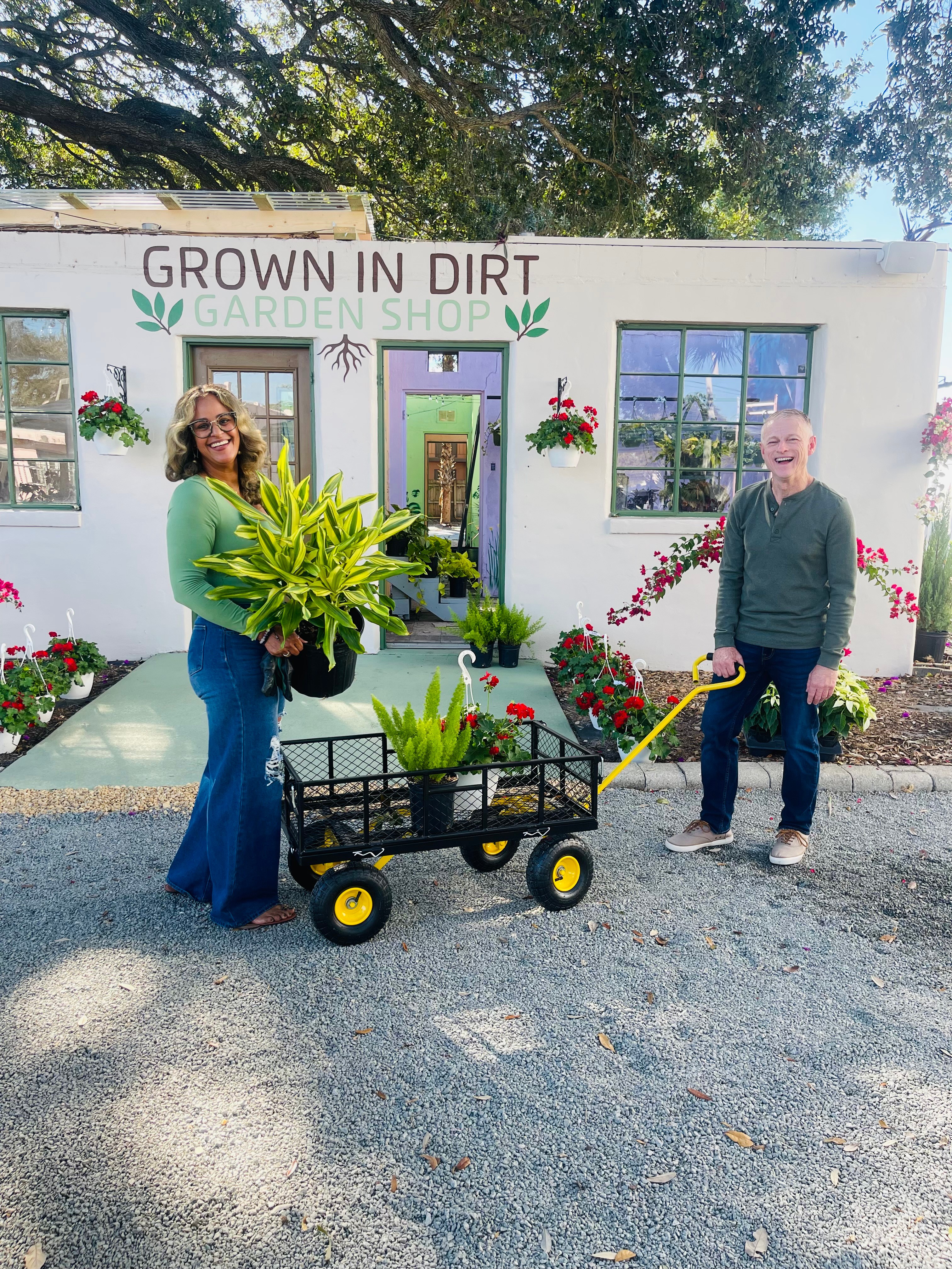 Two people stand outside a garden shop holding a large plant, with a wagon filled with plants between them, in front of a white building with flowers. Word sign reads 'GROWN IN DIRT GARDEN SHOP'.