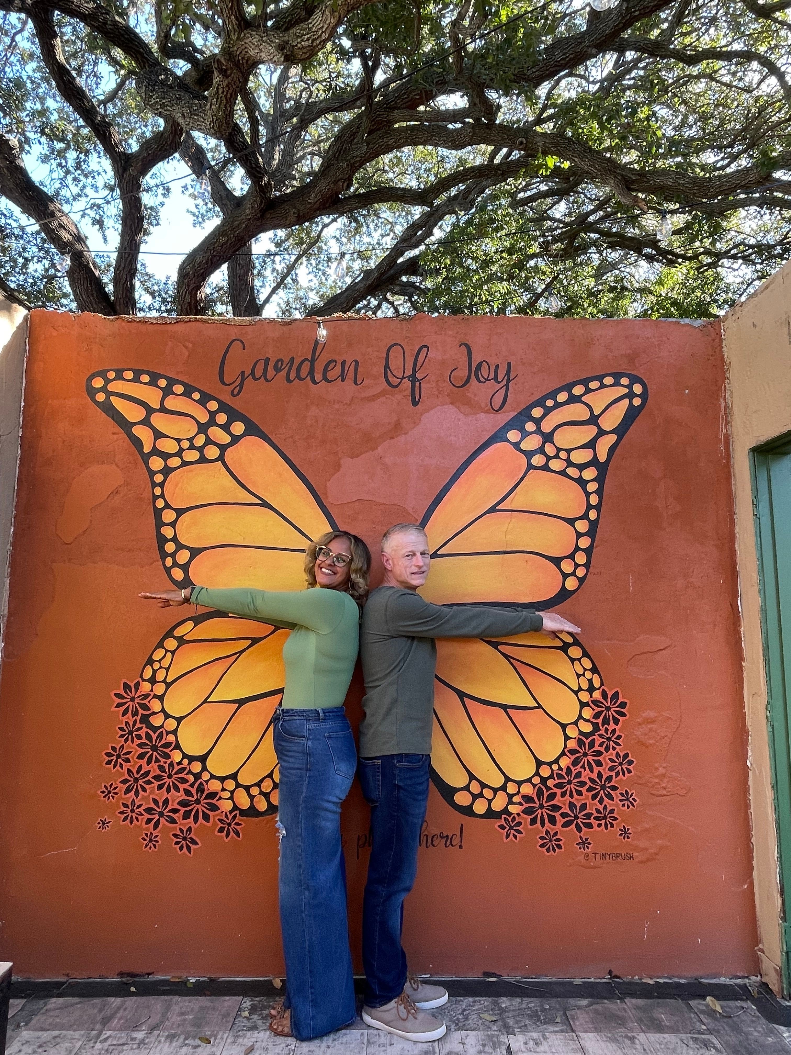 Person standing in front of a butterfly mural on a wall, with trees in the background.