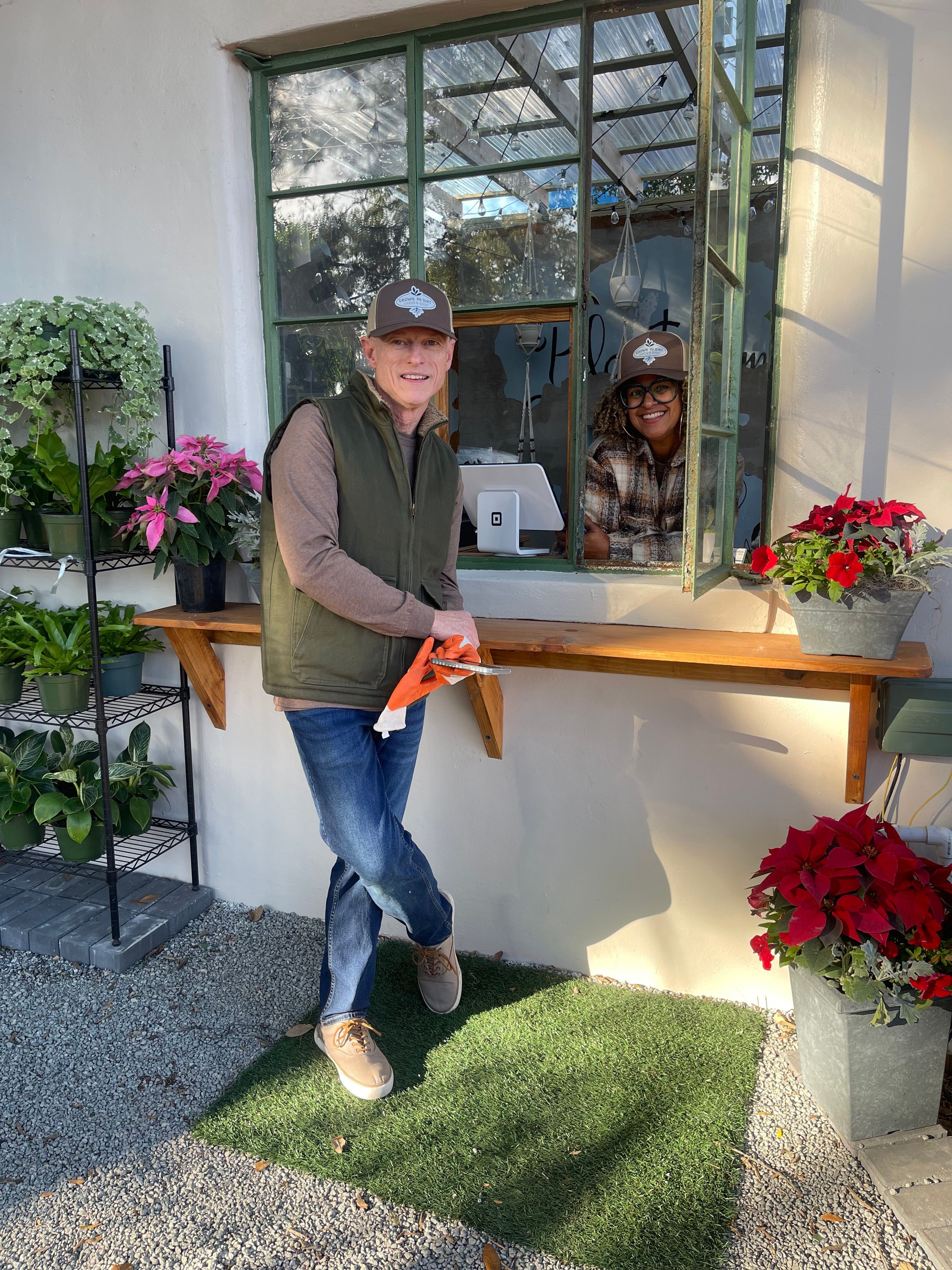 Person standing indoors near flowers and a glass window, wearing a pink cap, green vest, and jeans.