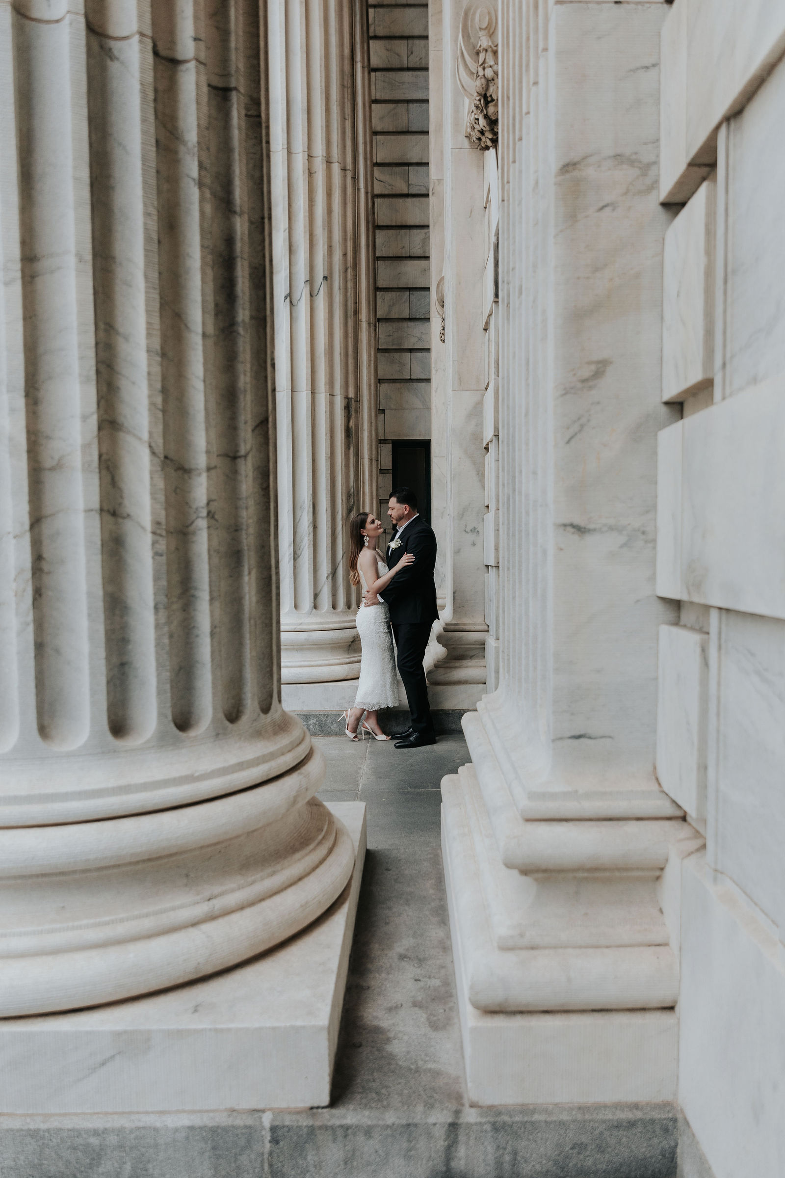 Couple standing between large classical columns, facing each other, in front of a stone building, with architectural details visible.