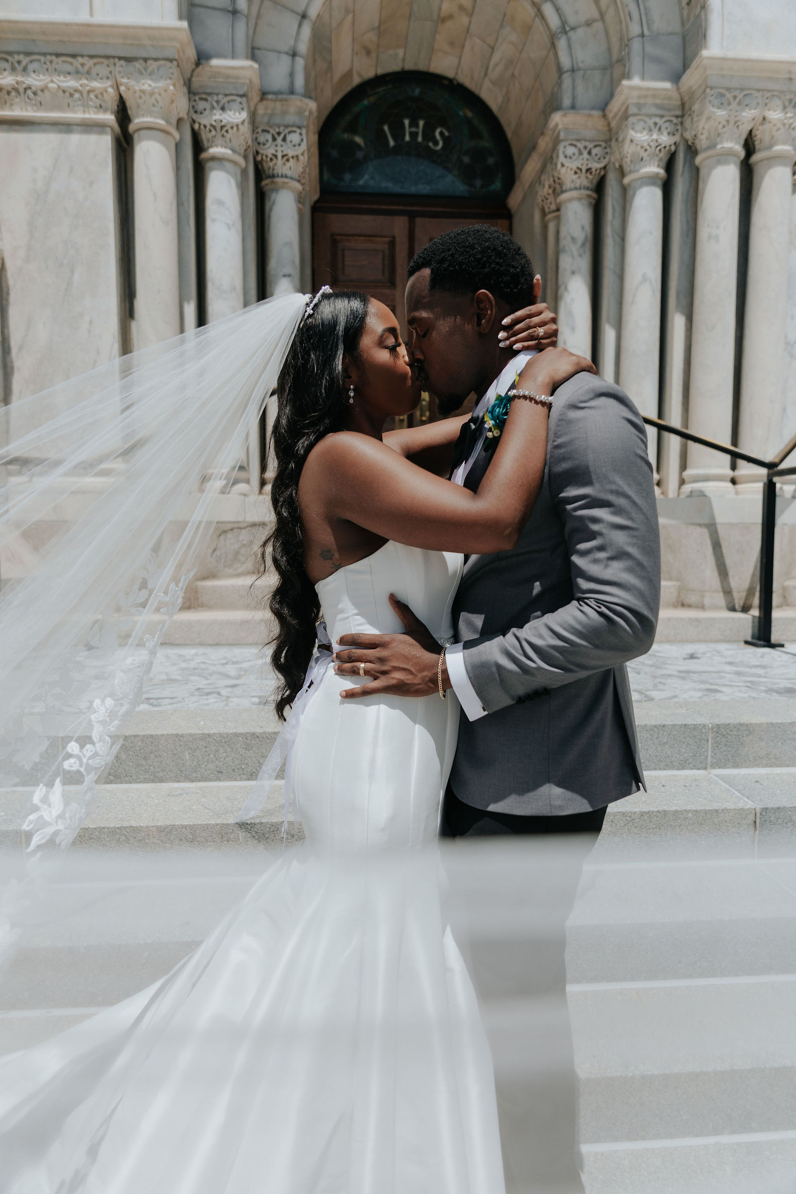 Couple kissing on steps outside a building, woman in wedding dress and veil, man in suit, stone columns in background.