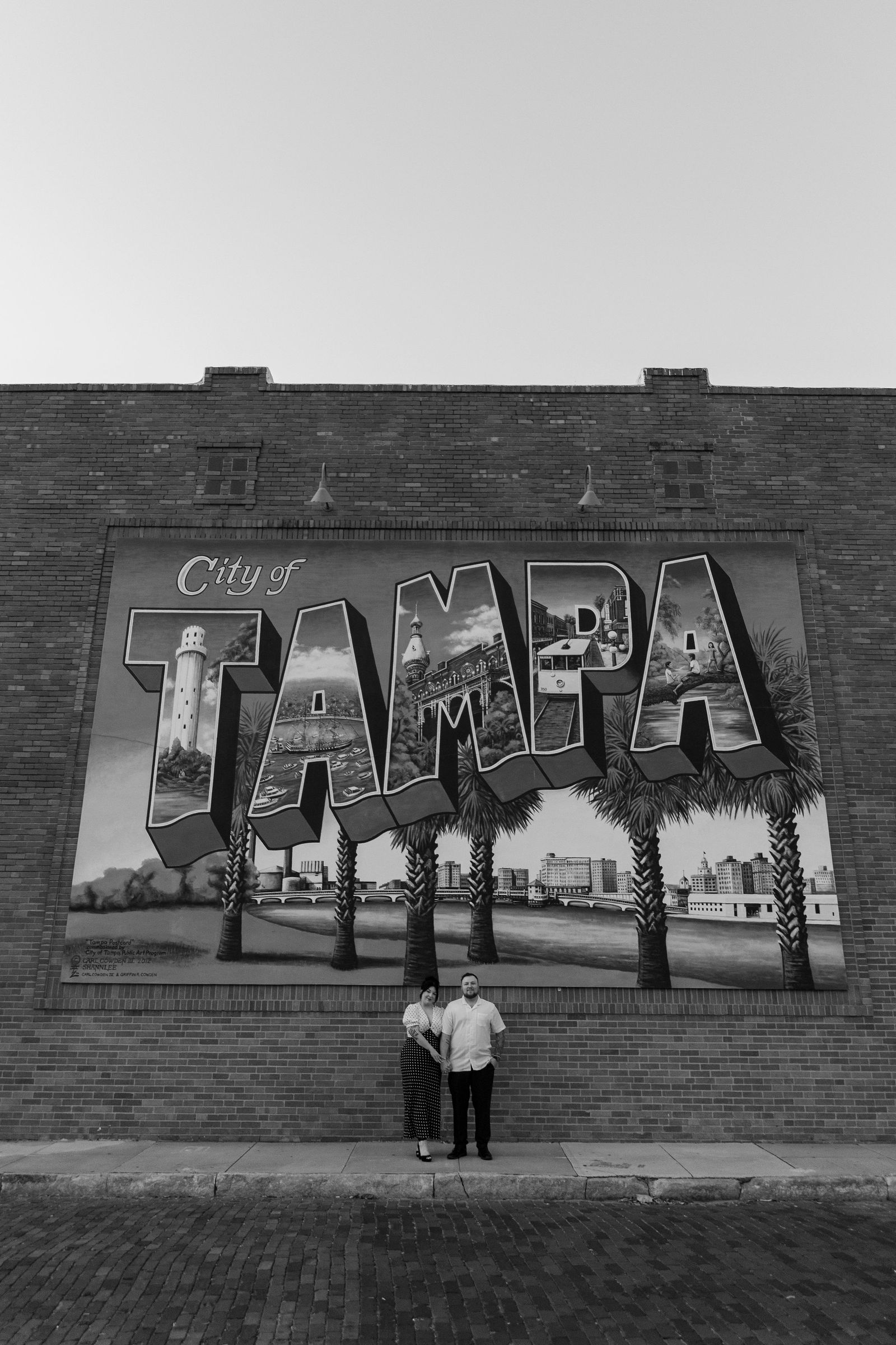 Two people stand in front of a large sign that reads 'City of TAMPA' with trees and a building behind.