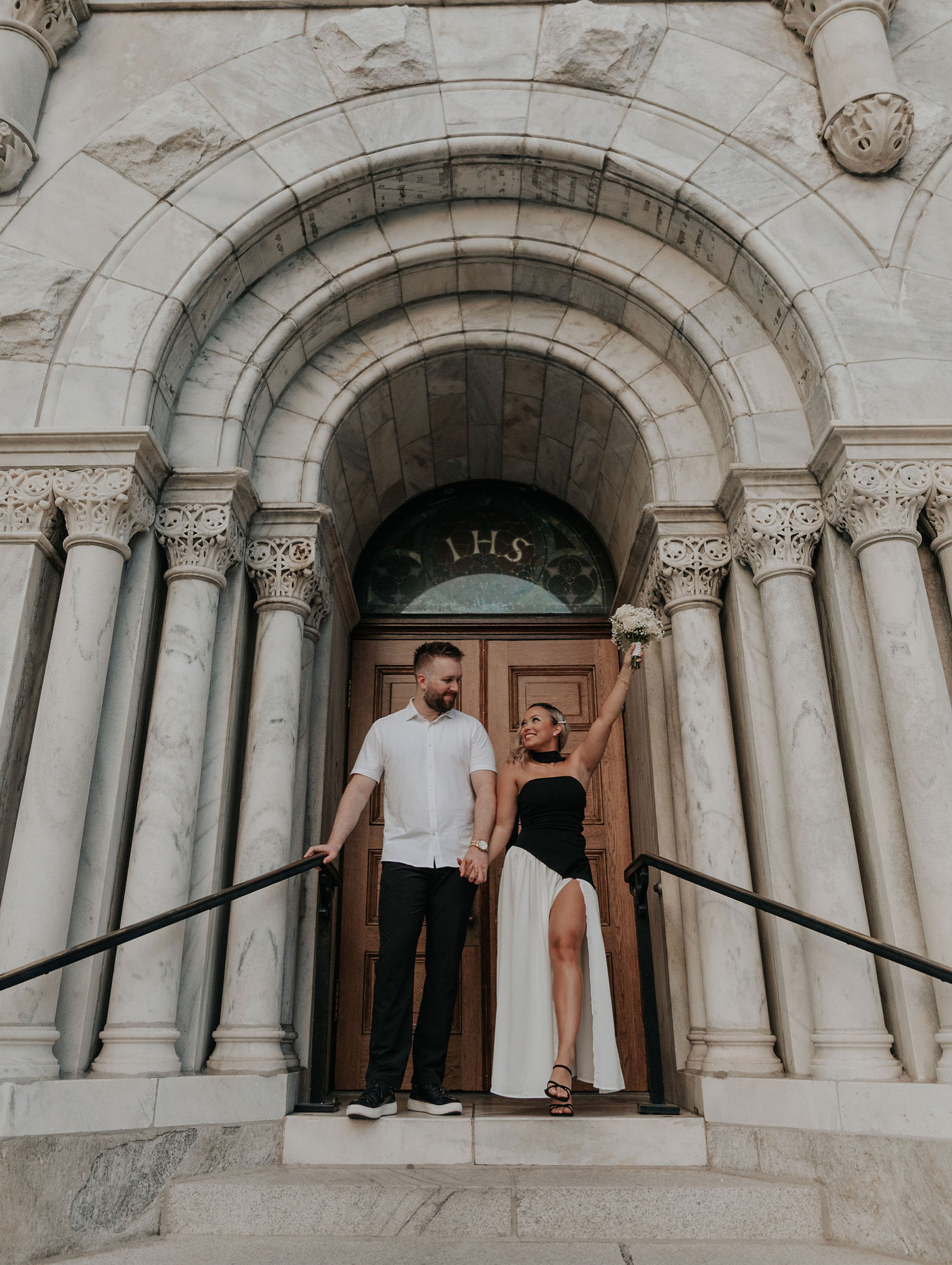 A man and woman stand in front of a large stone archway with columns, the woman is wearing a black dress with a high slit.