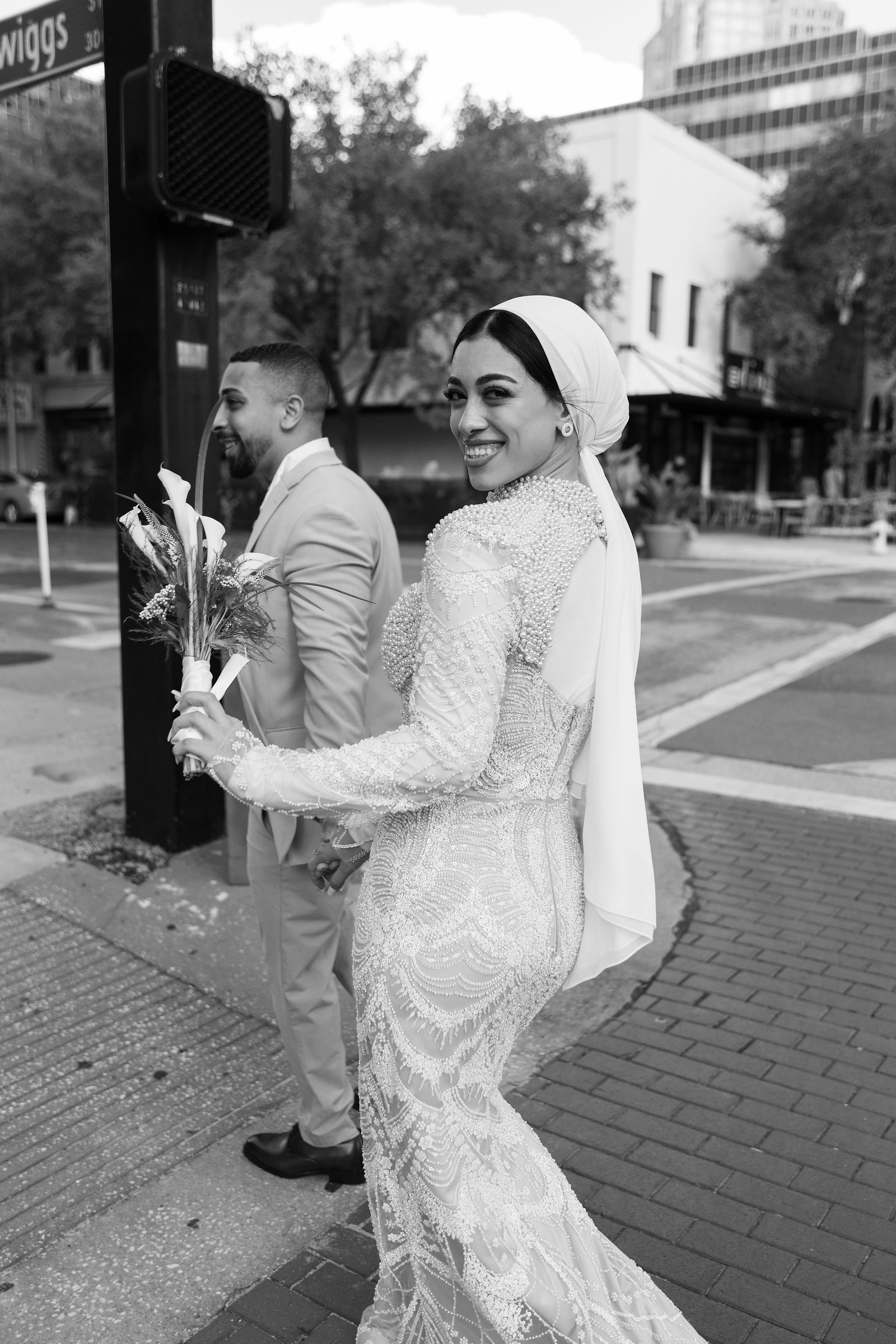Woman in wedding dress holding bouquet, smiling, on city sidewalk with man in suit behind her, black and white photo.