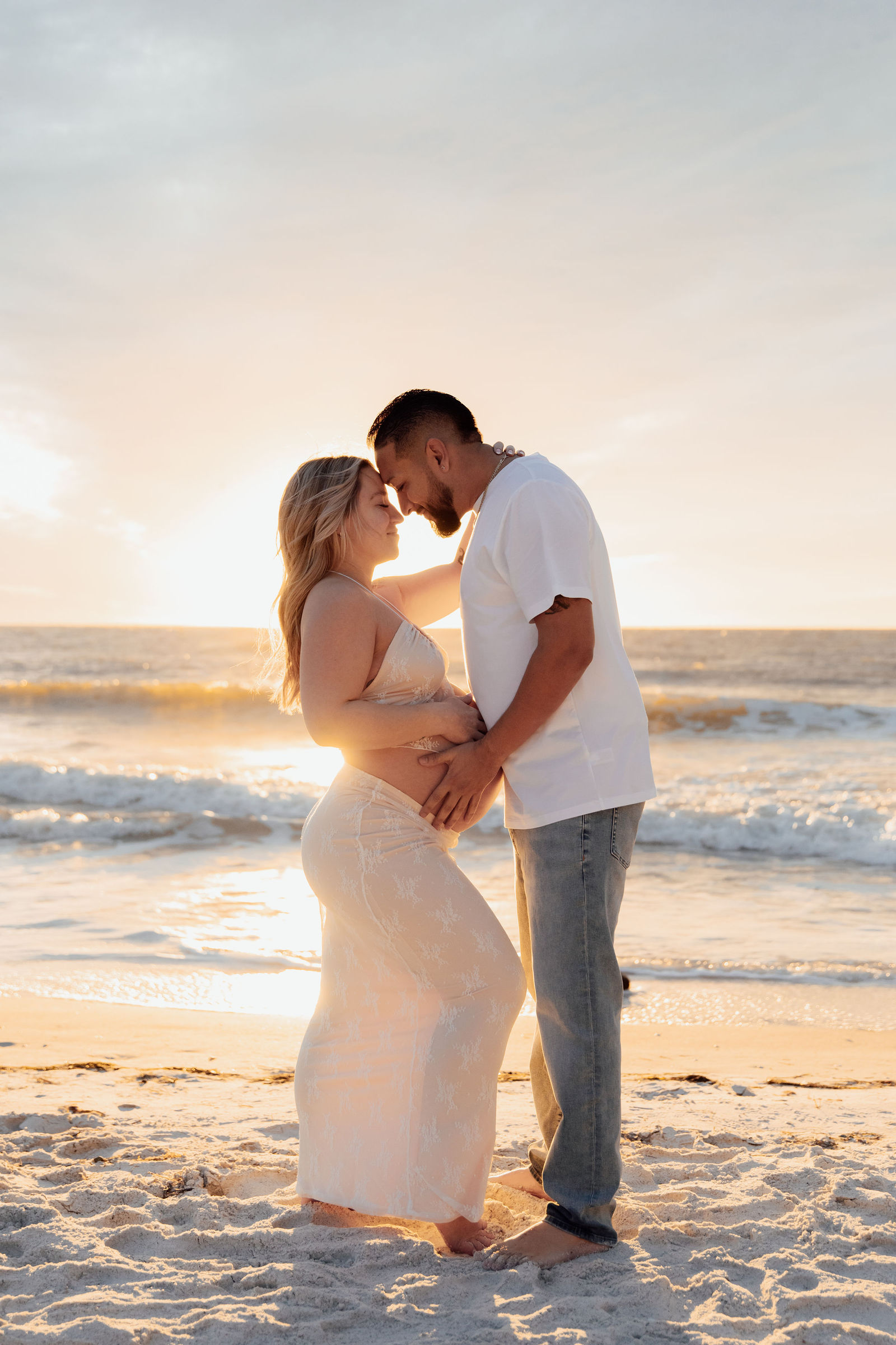 A couple stands close on the beach at sunset, facing each other and holding hands, with the ocean in the background.
