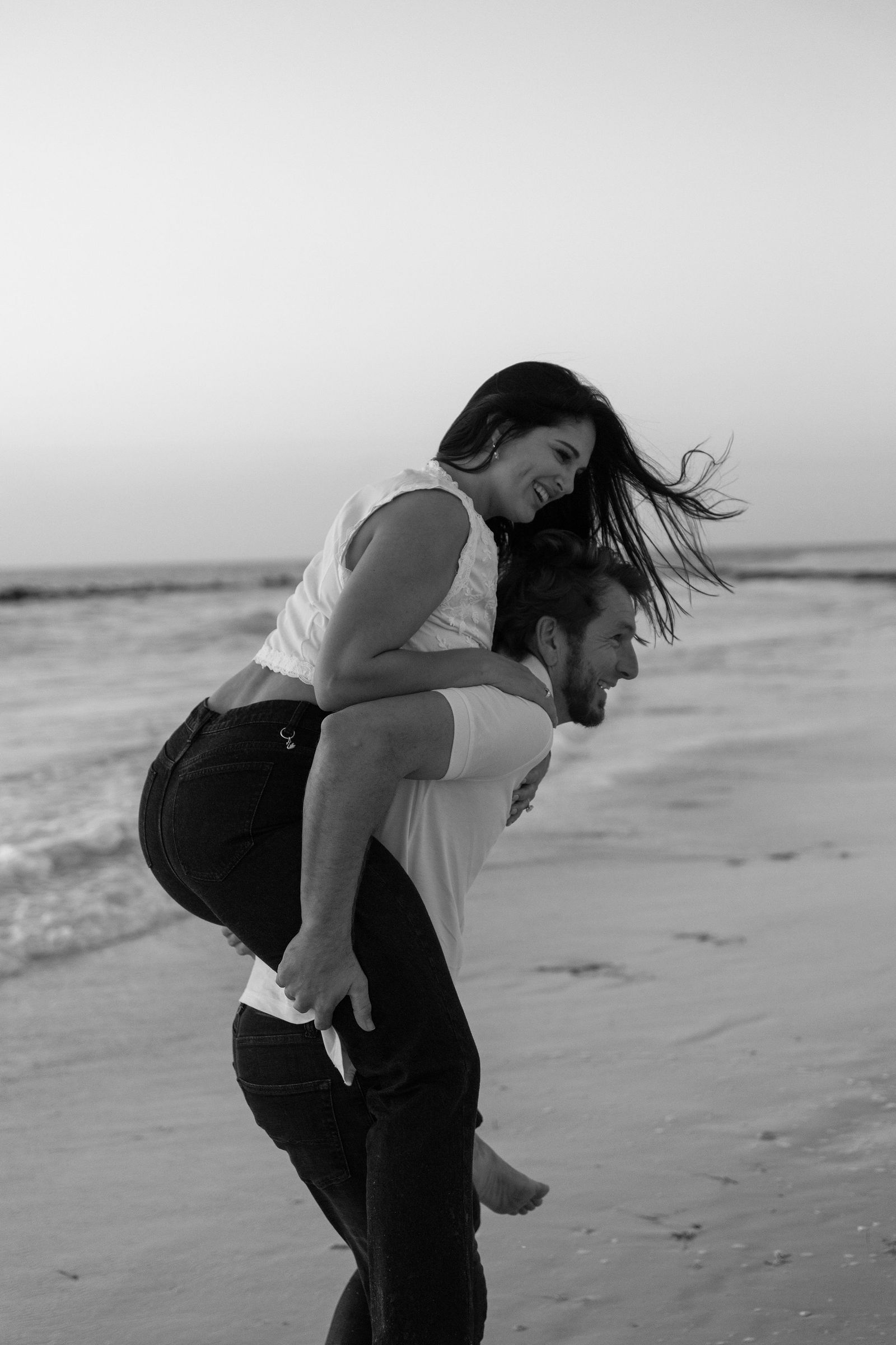 A couple on the beach, the woman on the man's back, smiling and enjoying the moment, with wind blowing her hair.