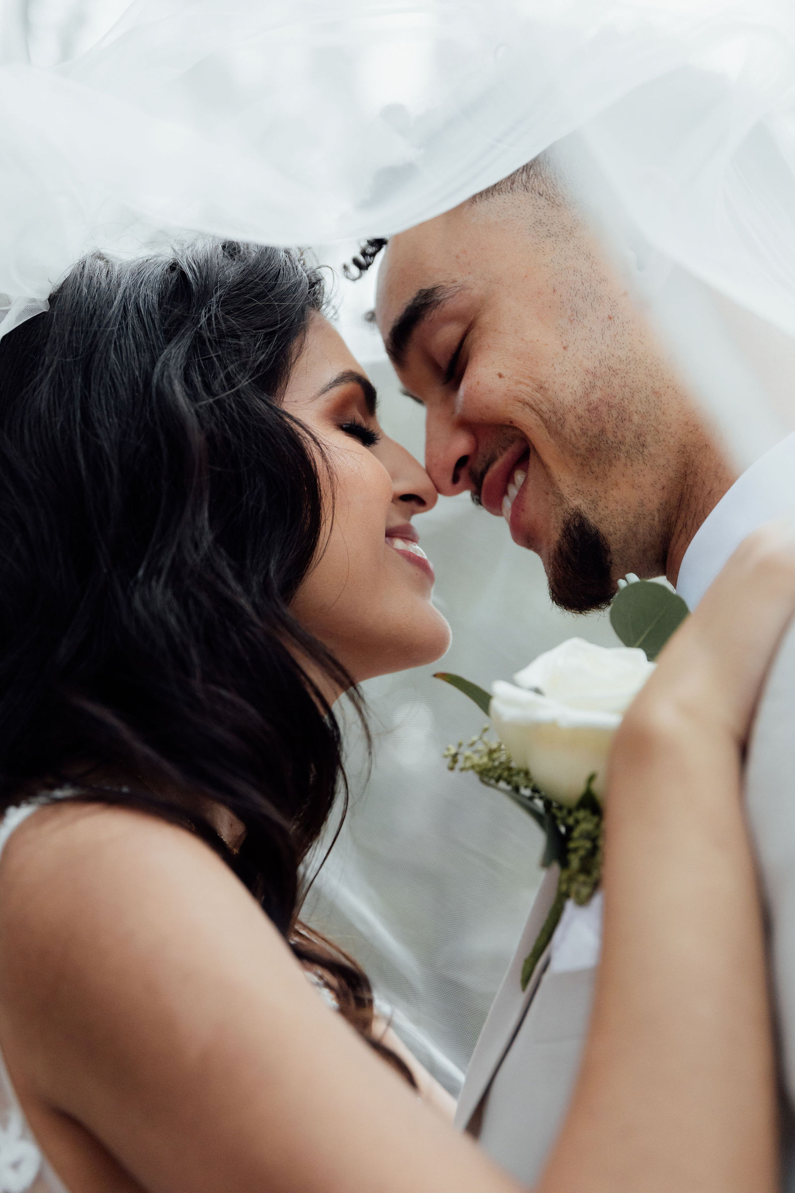Close-up of a couple smiling with foreheads touching, under a white veil, holding a white rose bouquet.