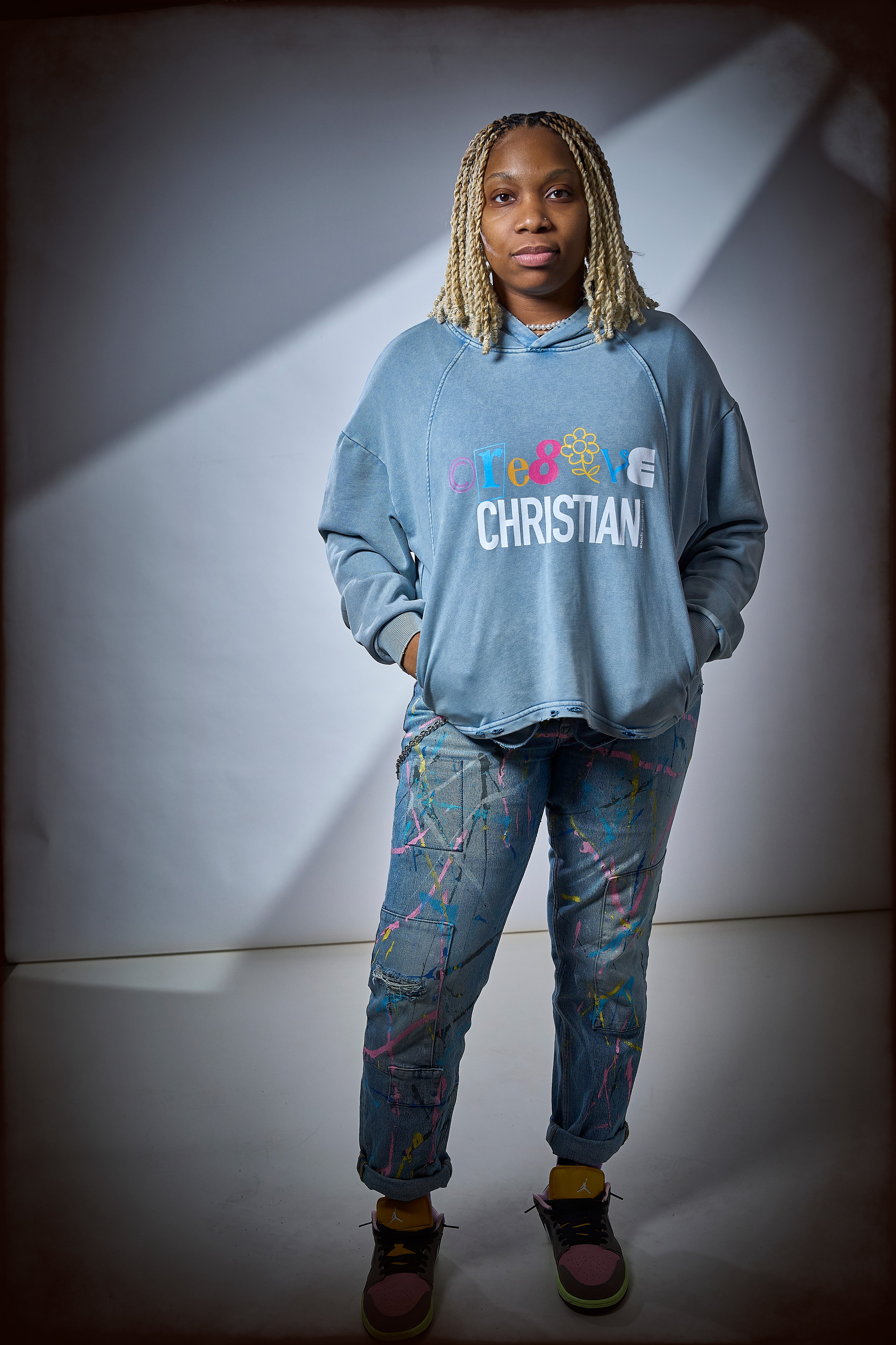 Young woman with blonde dreadlocks wearing a blue Christian sweatshirt and patterned pants, standing against a plain background.