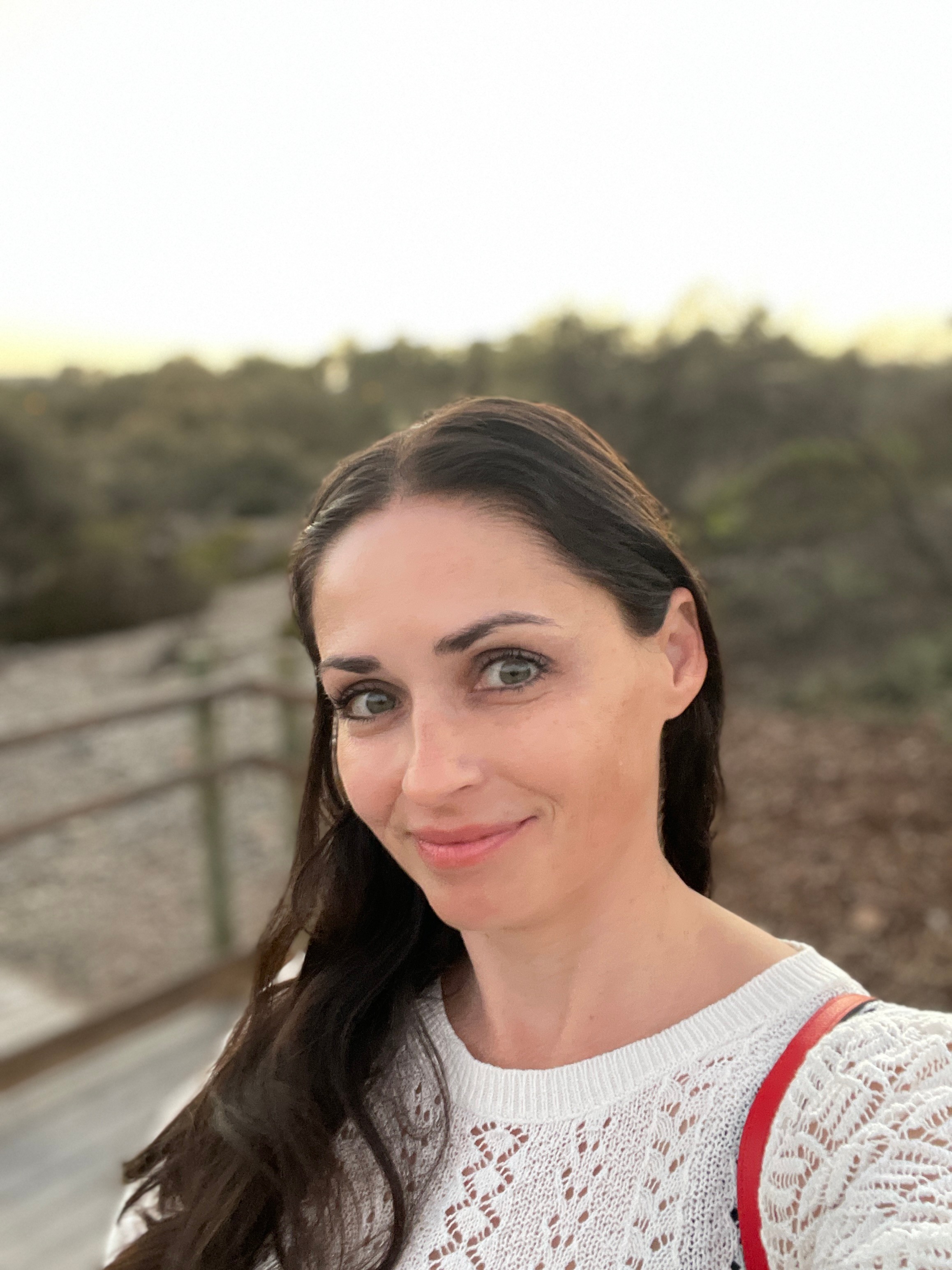 Woman with long dark hair smiling outdoors, wearing a white knit sweater and red strap, landscape background.