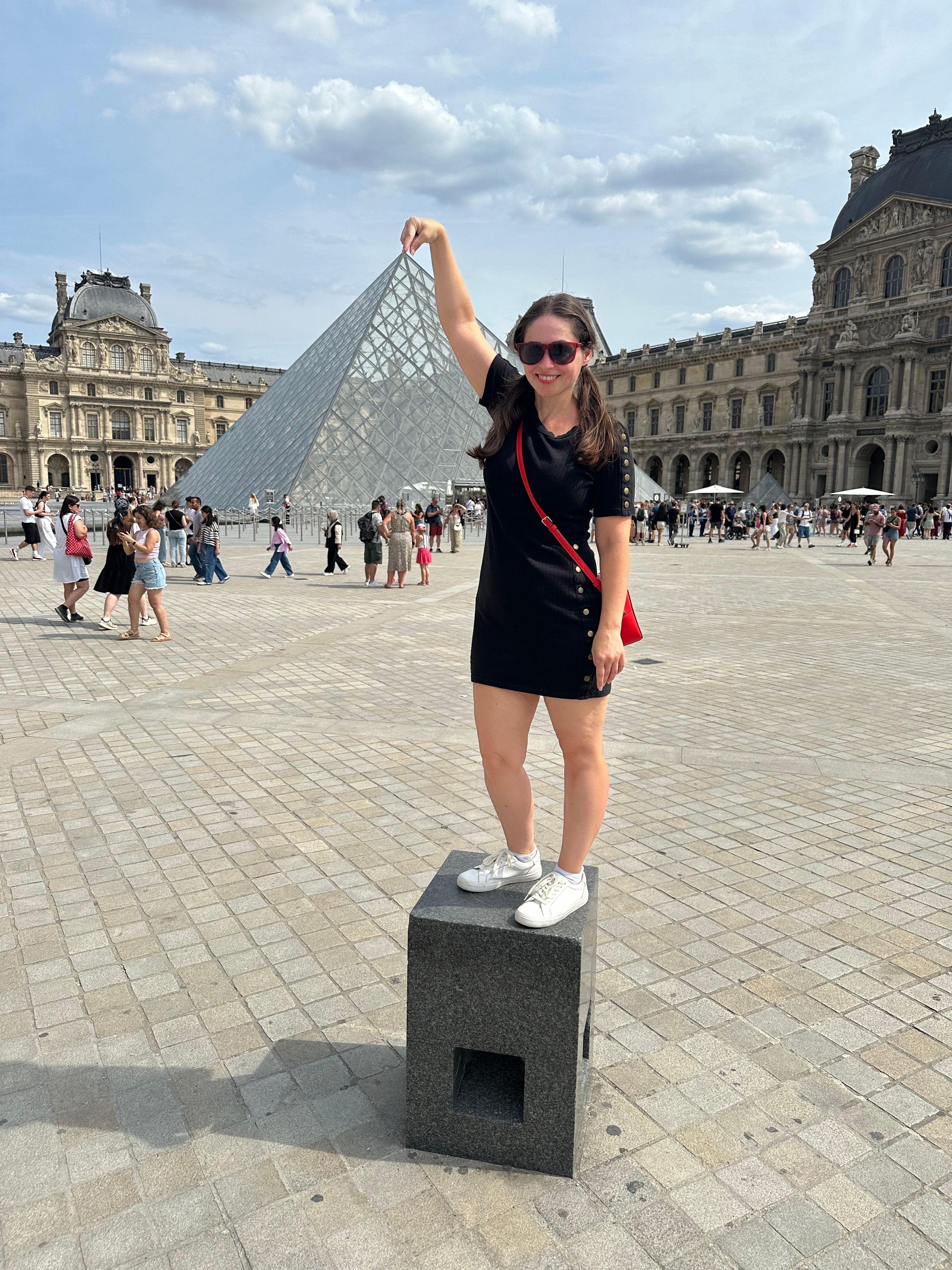 Person balancing on a black box in a public square with historic buildings and crowds, under a partly cloudy sky.