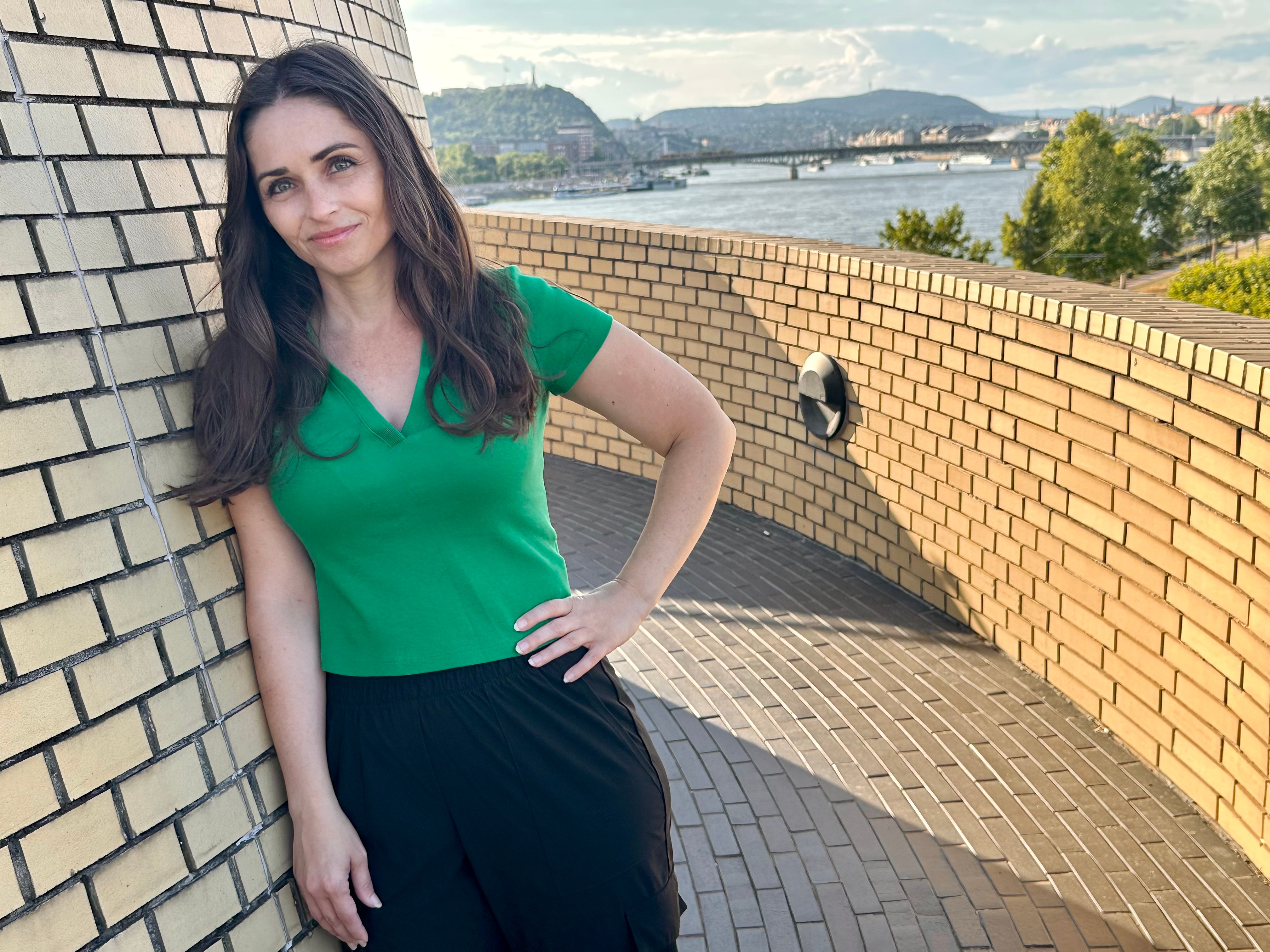 Young woman with long dark hair in a green top and black pants leaning against a brick wall outdoors, with a river and cityscape in background.