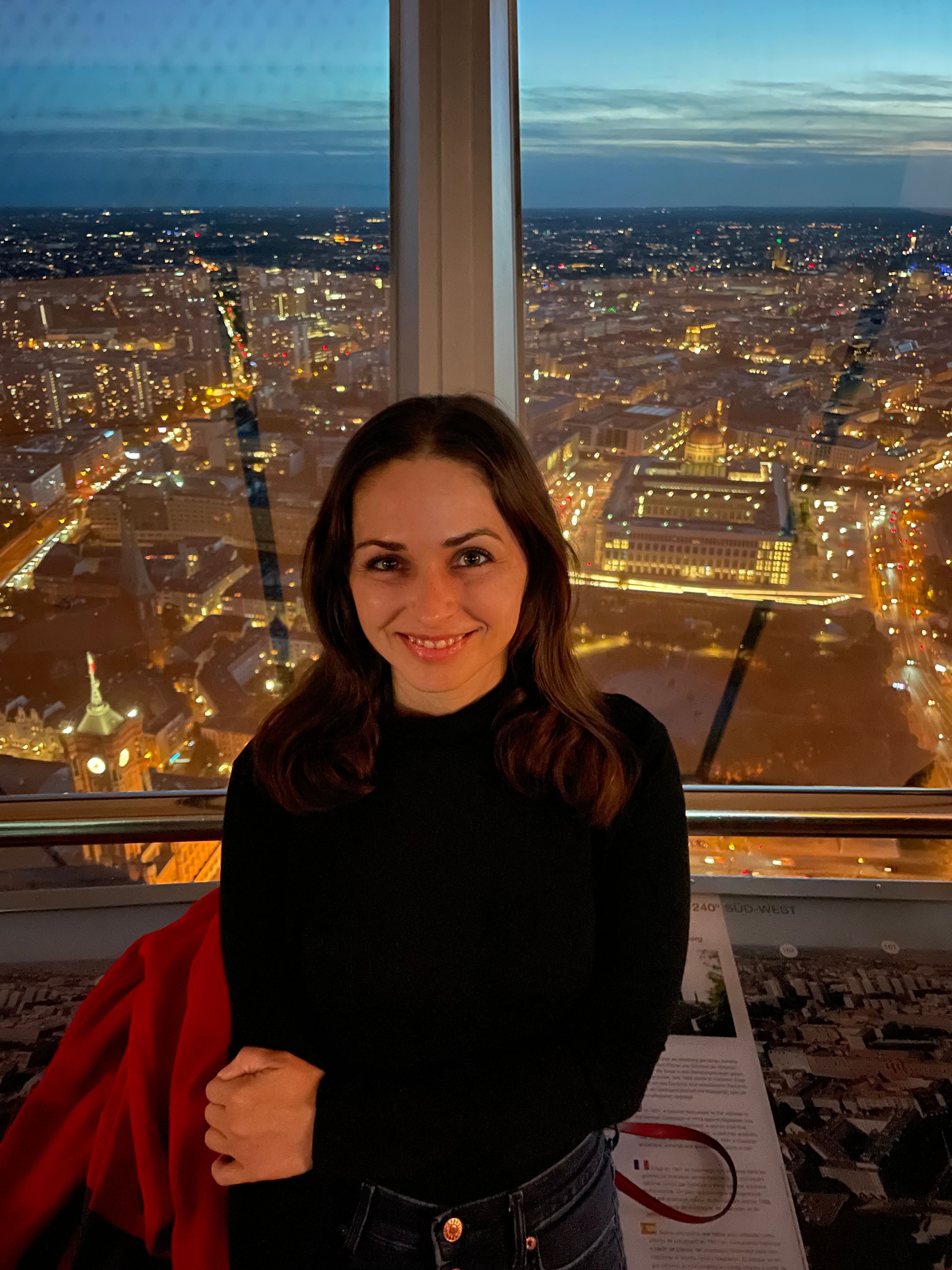 Woman with dark hair in a black top, standing indoors with city lights outside the window at night.