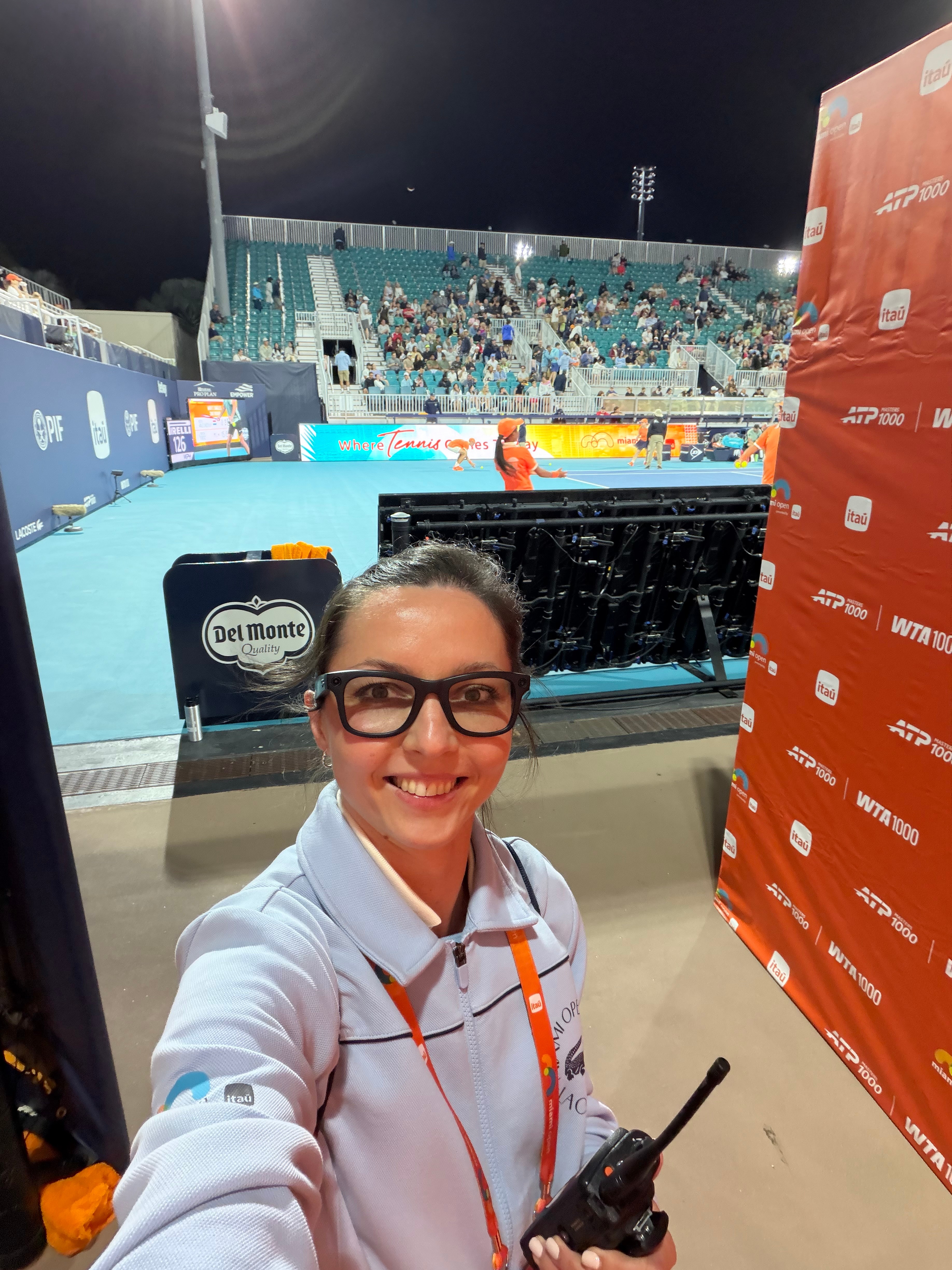 Smiling woman with glasses taking a selfie at a sports event, with a tennis court and spectators in the background.