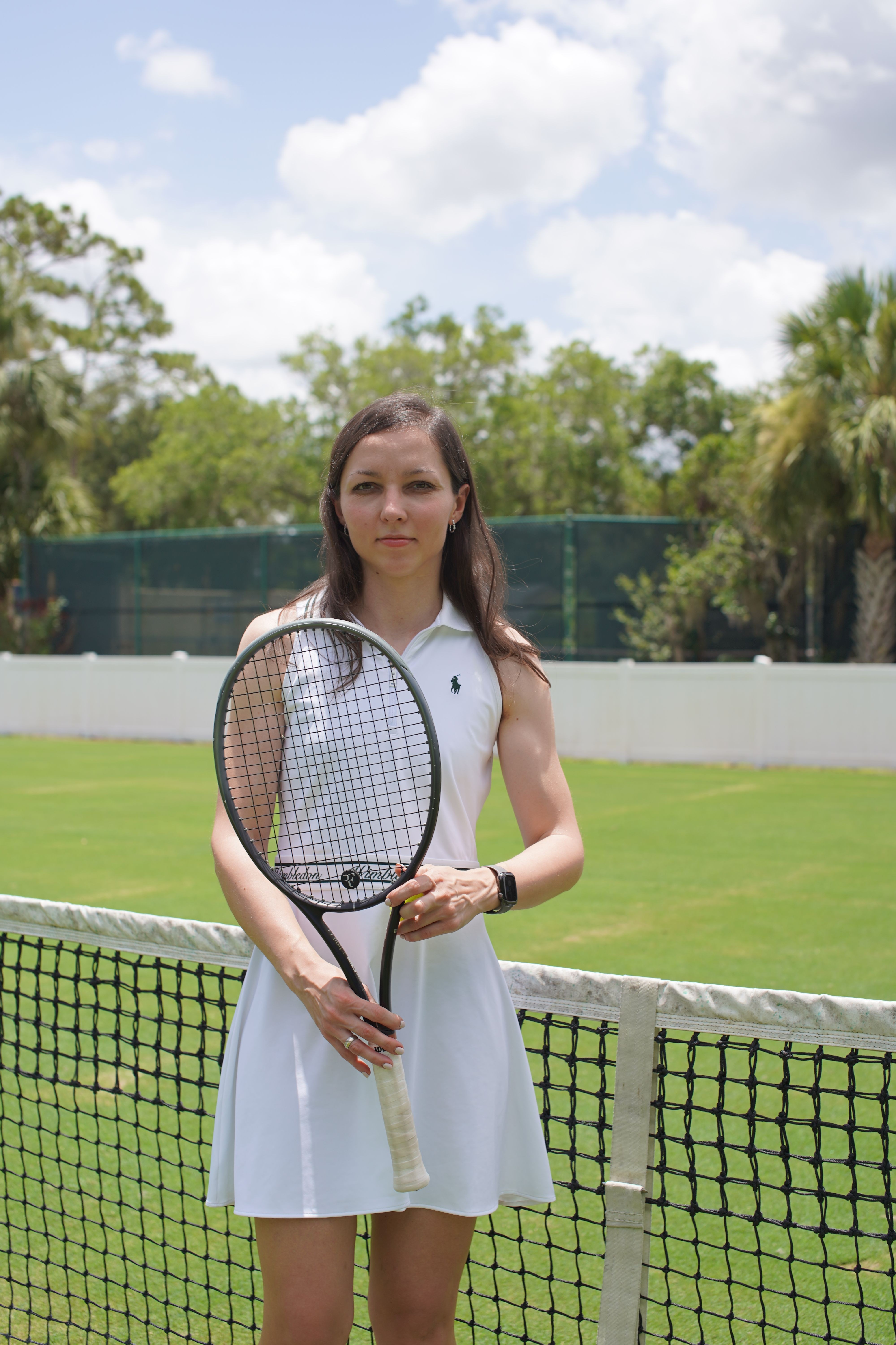 A woman in a white tennis dress holding a tennis racket on a tennis court with green grass and trees in the background.