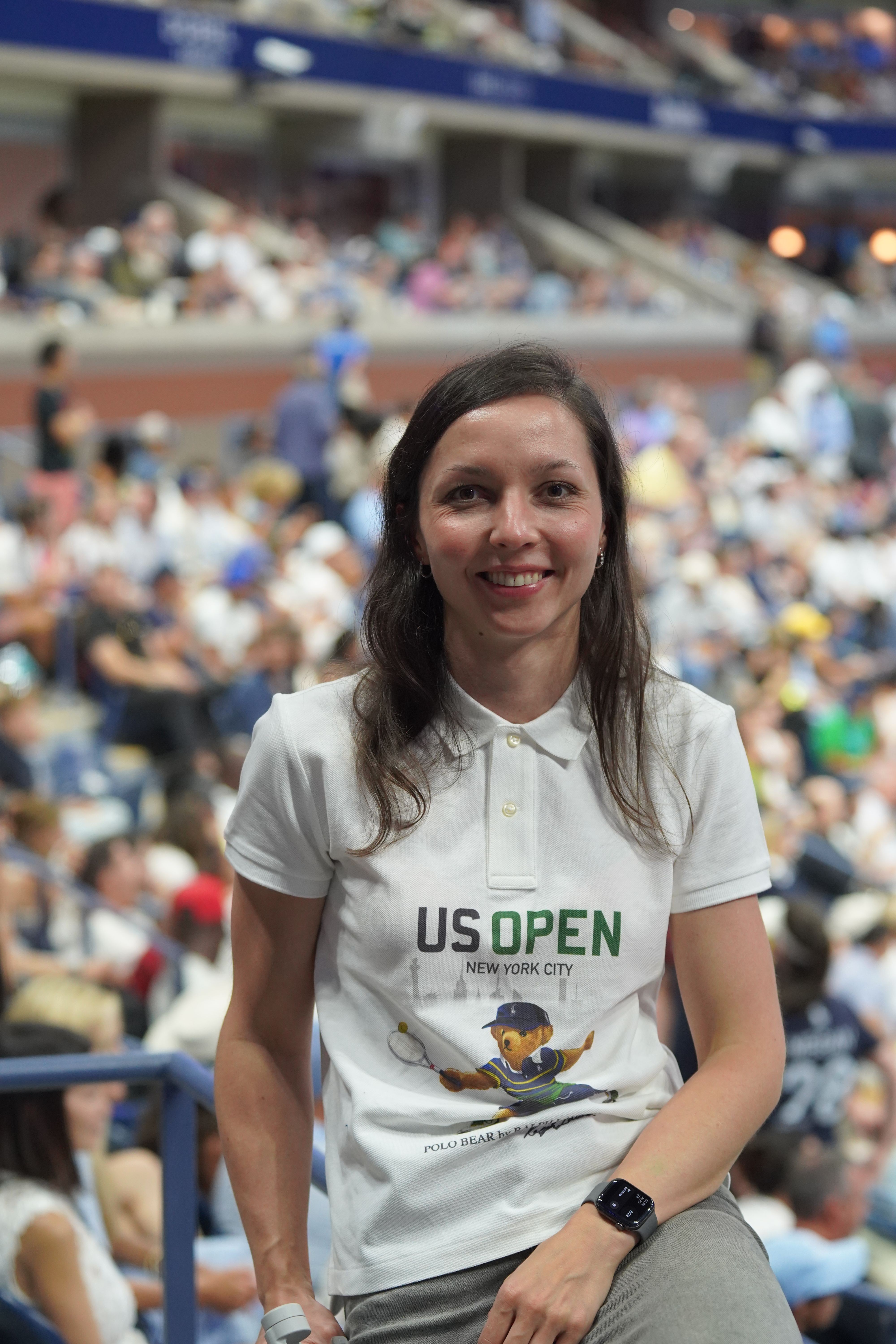 Woman wearing a white US Open t-shirt sitting in a crowded outdoor area, smiling, with people in the background.