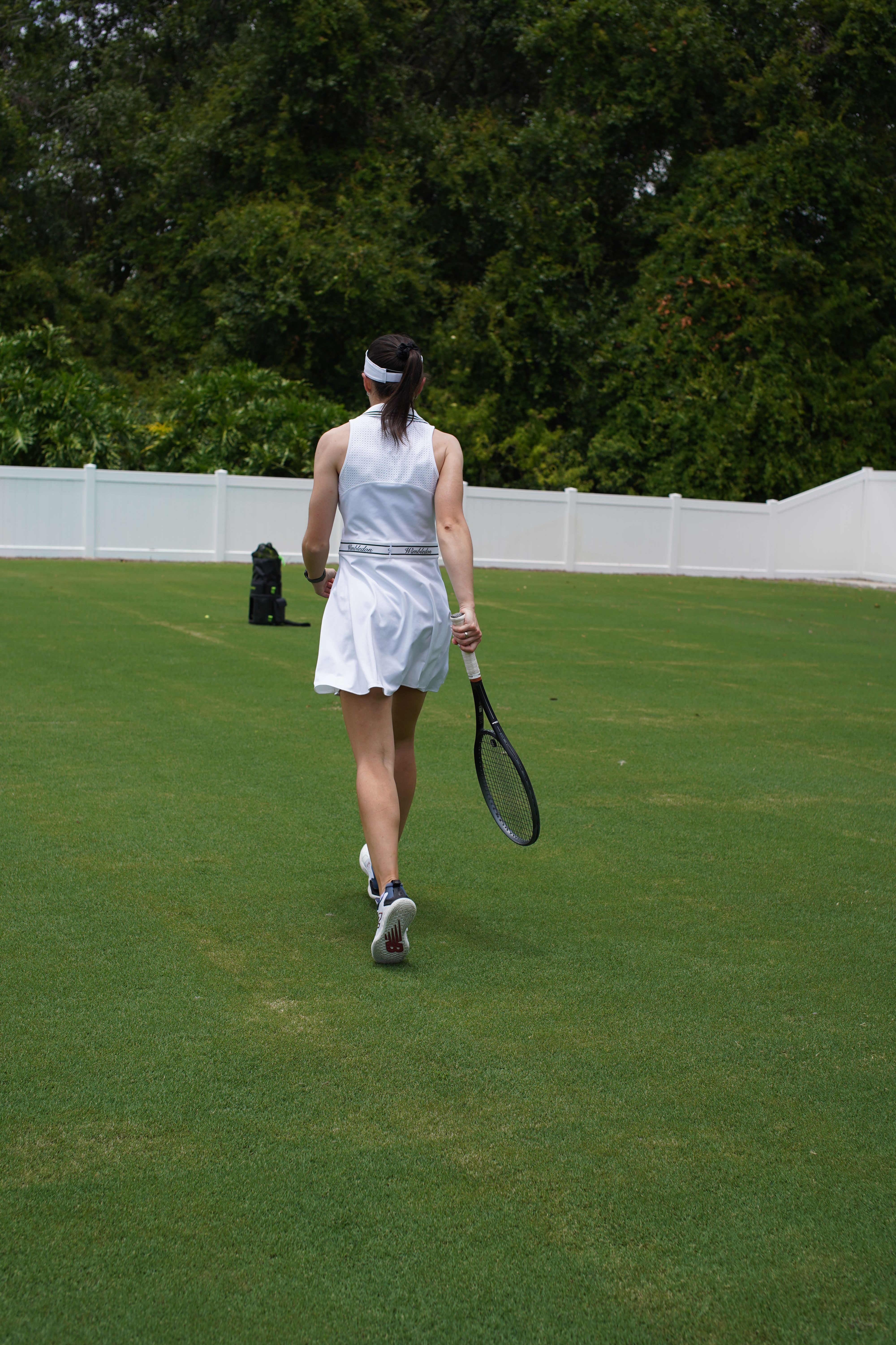 A person in a white dress holding a tennis racket on a grass court, with trees and a white fence in the background.