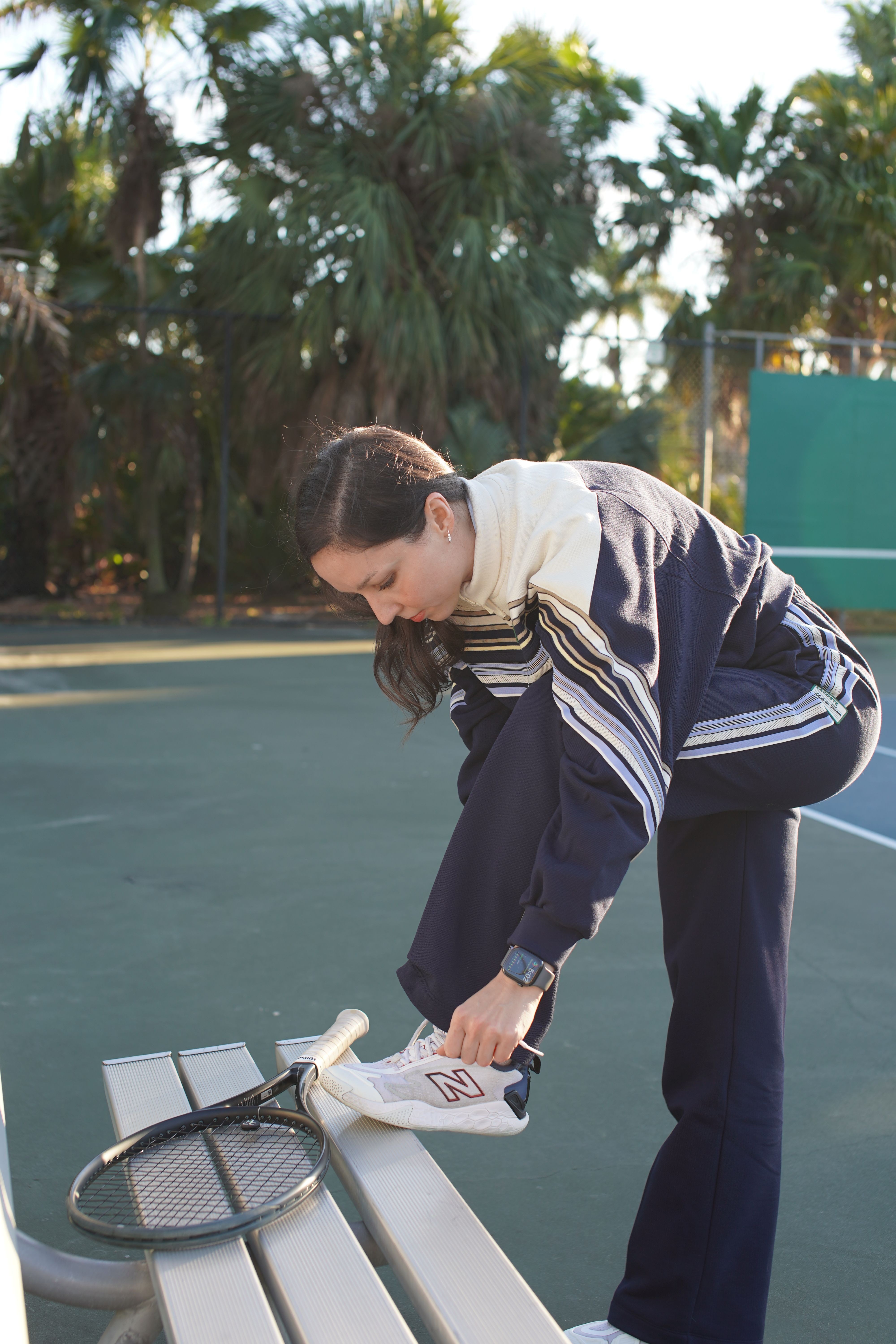 Person sitting on a bench holding a tennis racket, outdoors with trees in the background.