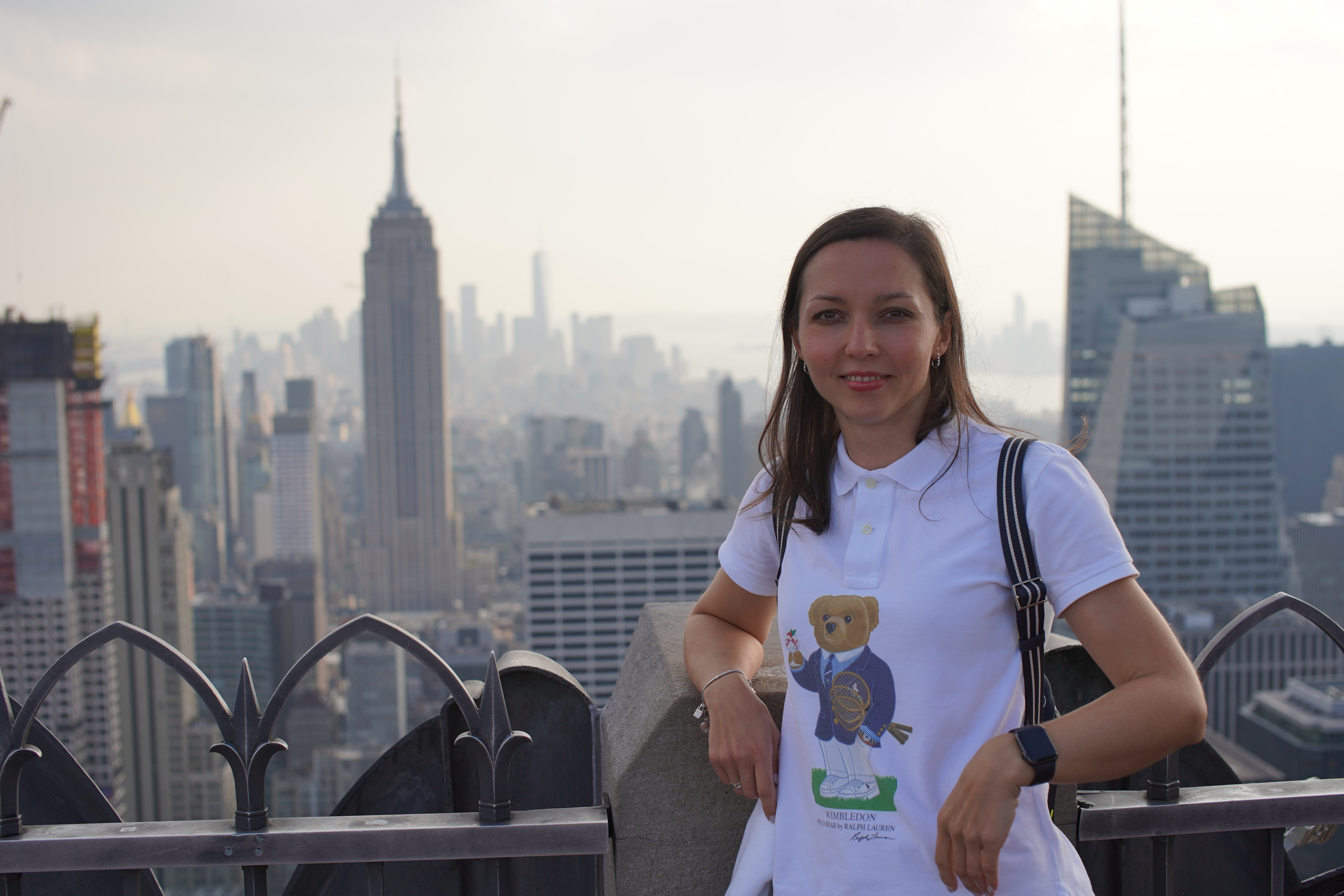 Young woman standing on a rooftop with New York City skyline in background, wearing a white T-shirt with a bear graphic, smiling.