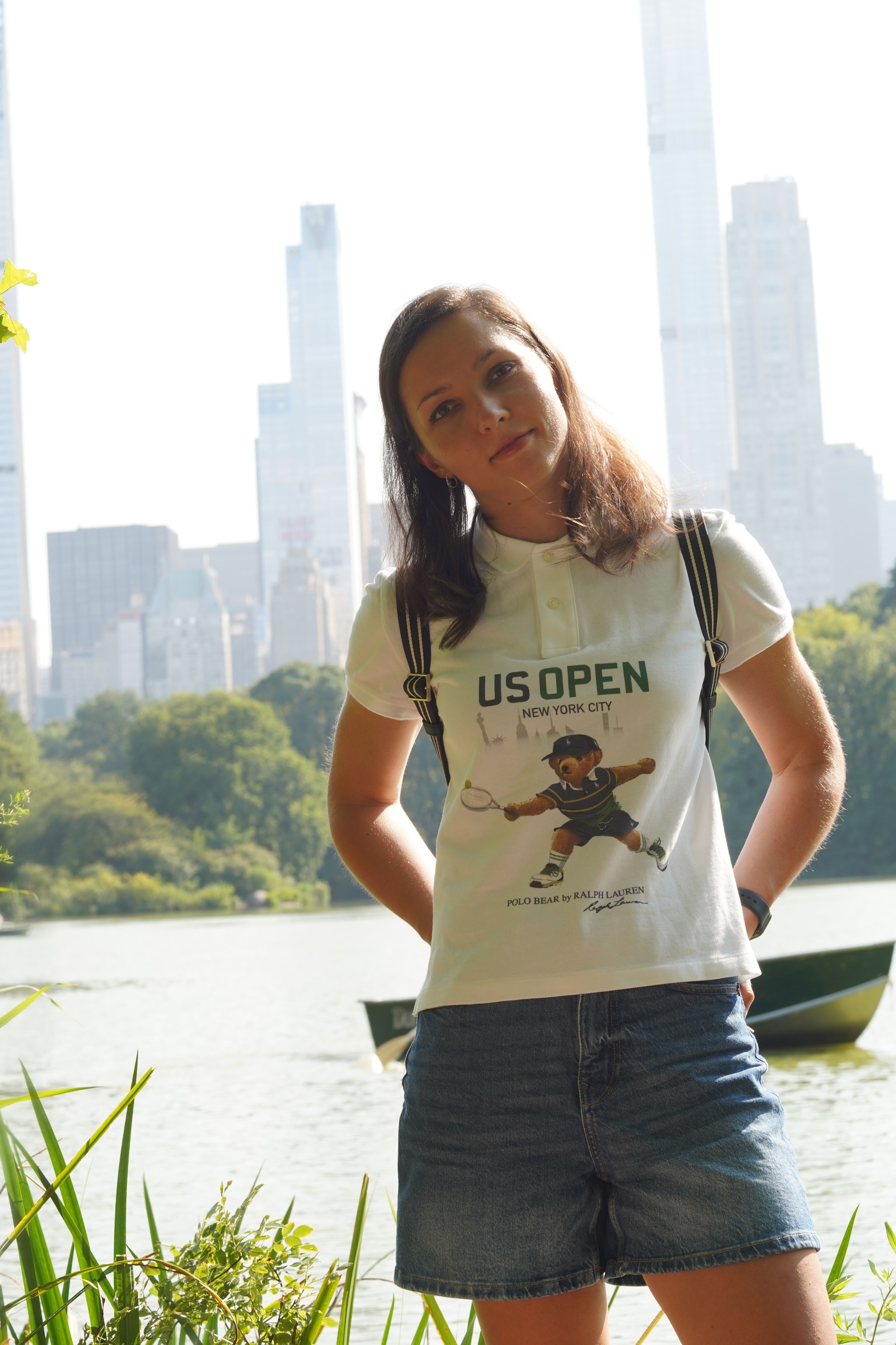 Woman standing outdoors near water with city skyline in background, wearing a graphic t-shirt and jeans, smiling.