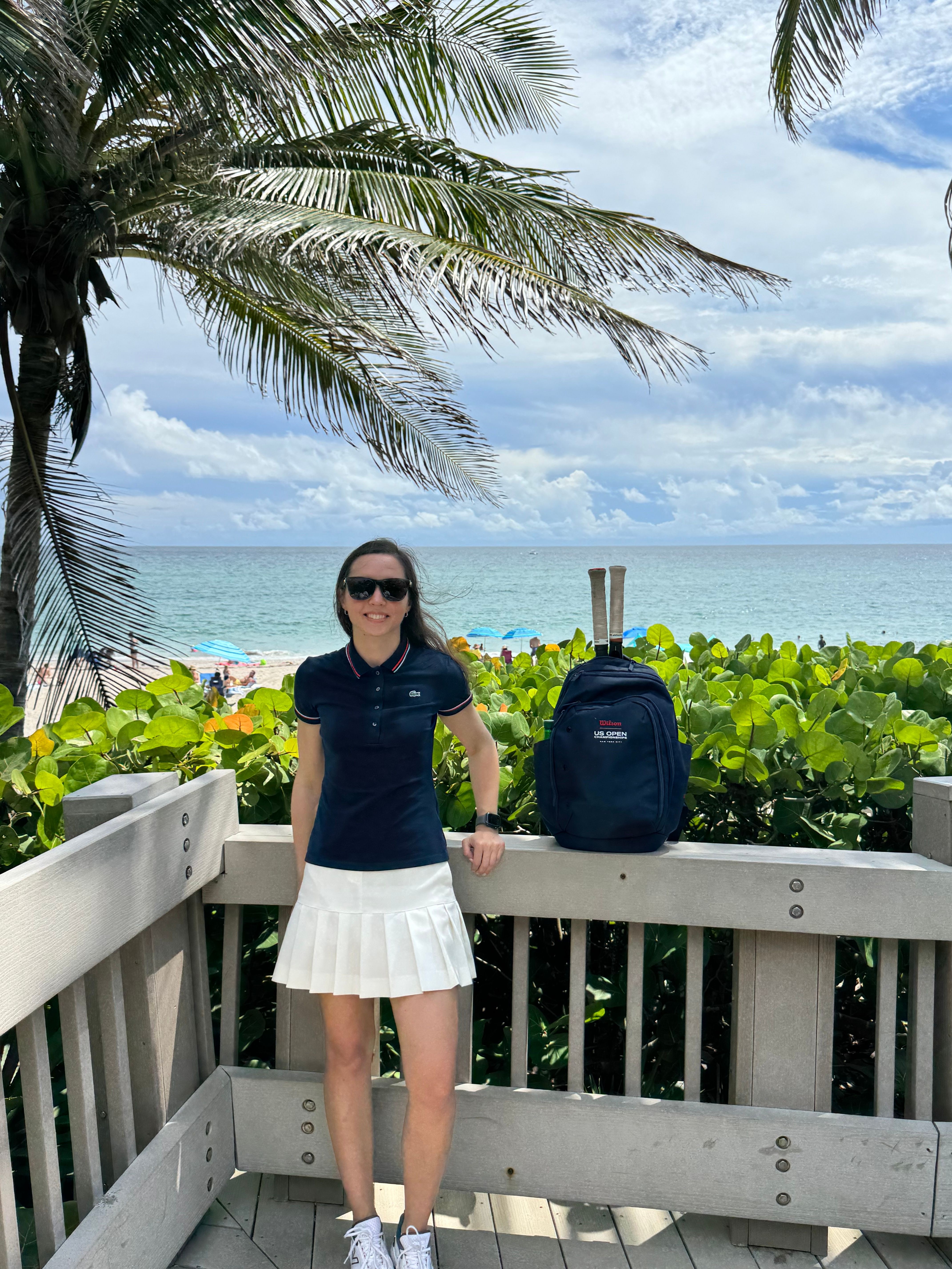 Woman standing on a wooden deck near a palm tree, with ocean and sky in the background, carrying a backpack.