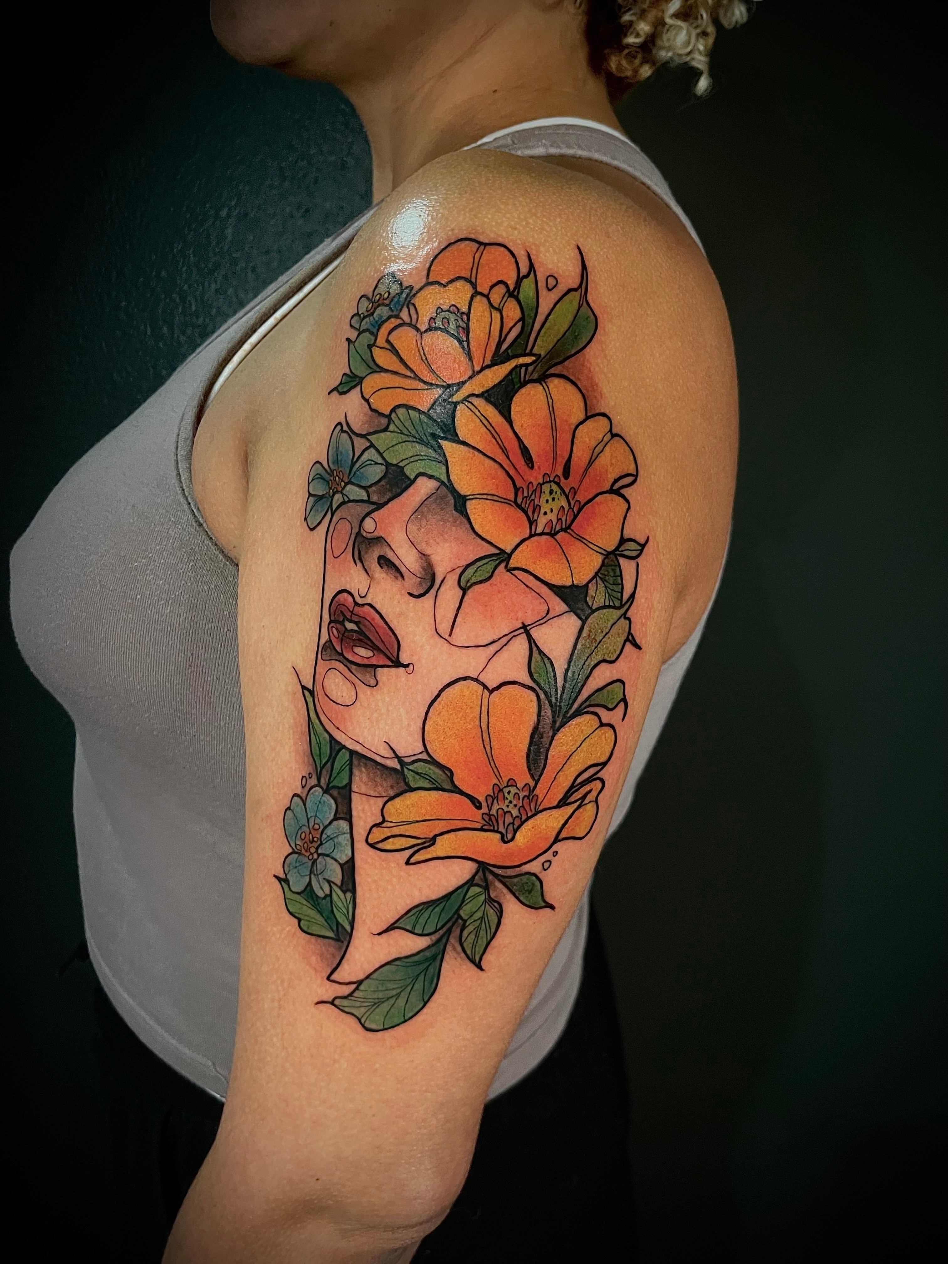 Tattoo of a woman's face surrounded by orange and blue flowers on upper arm, with dark background.