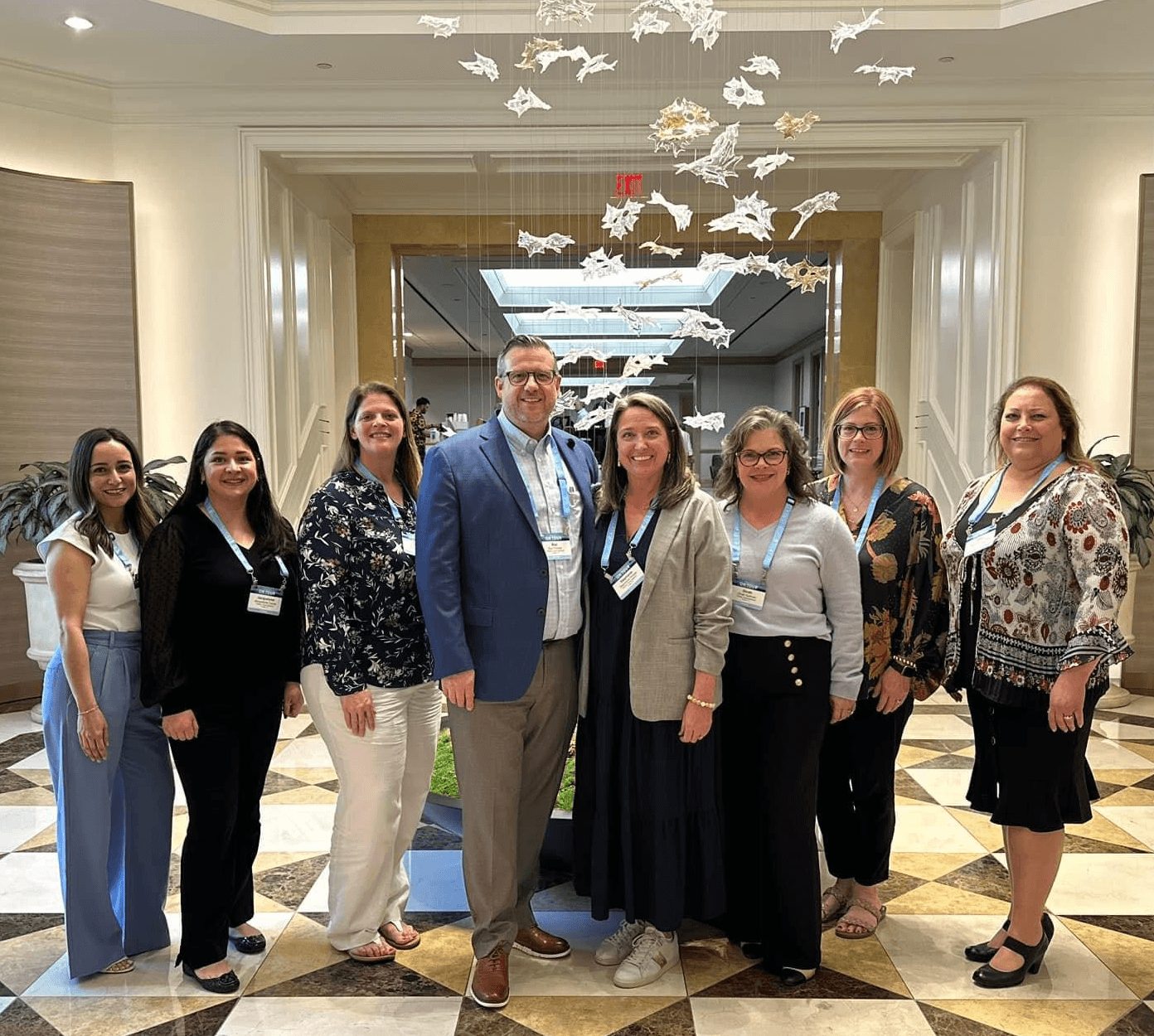 Group of eight people standing in a hotel lobby with a fish sculpture overhead, smiling for the photo.