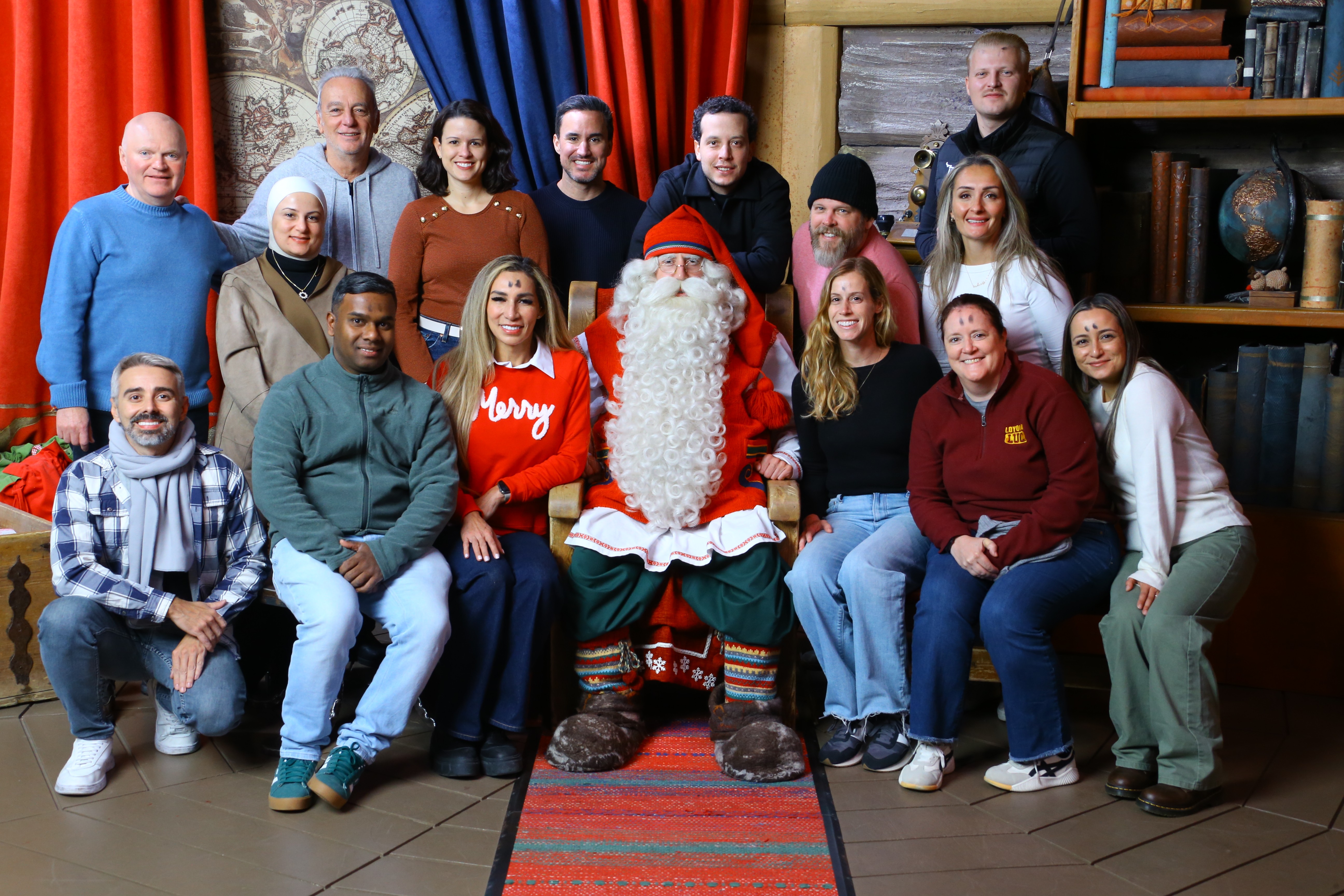 Group of people with Santa Claus in a cozy room, some smiling, some sitting, some standing, festive atmosphere.