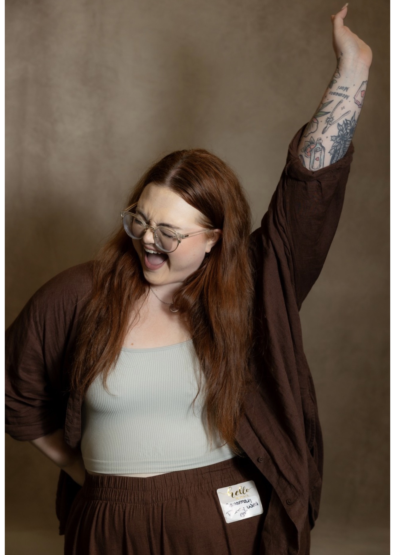 Woman with long red hair, glasses, and tattoos, smiling and raising her right arm, wearing a white top and brown cardigan.