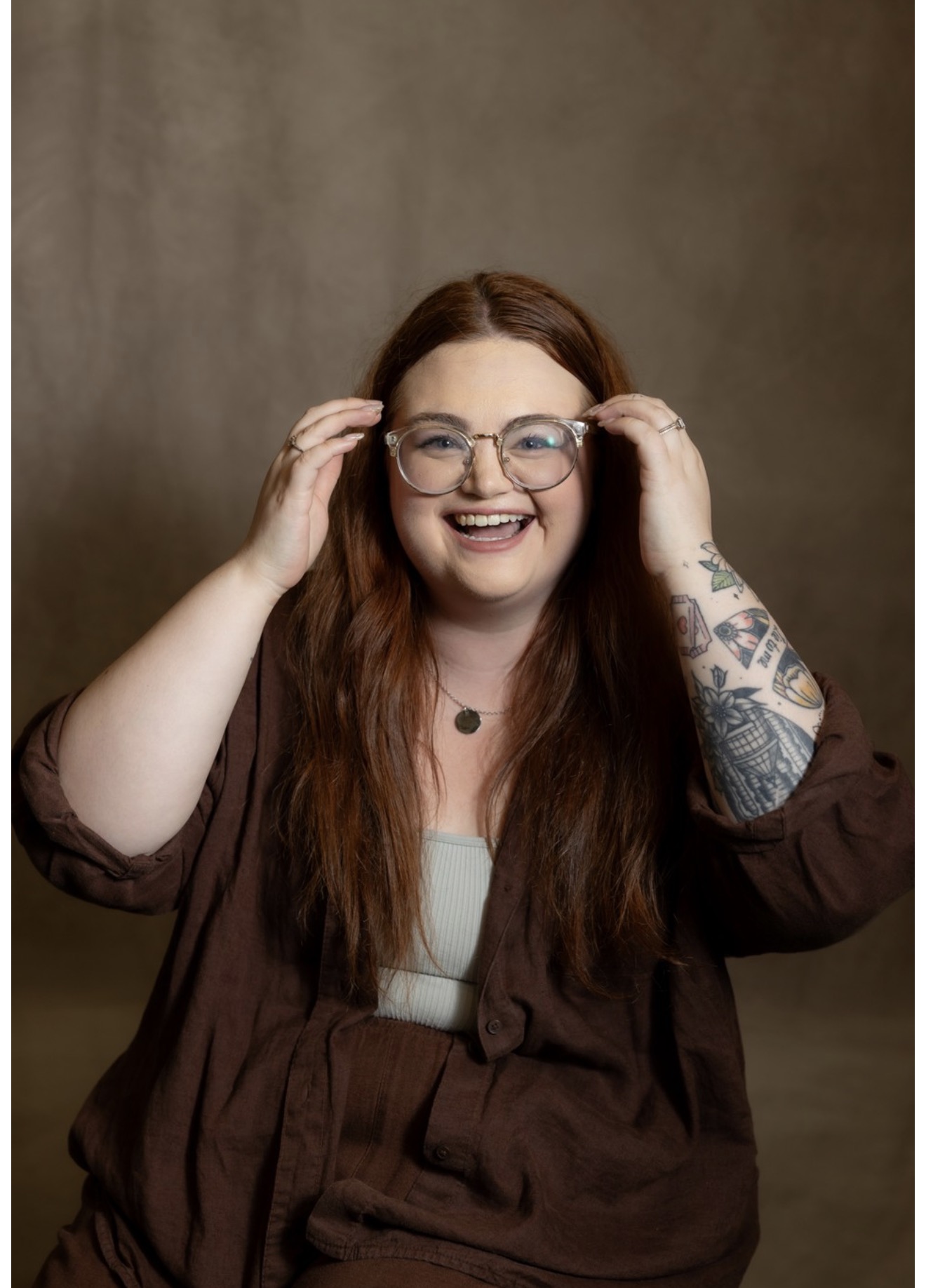 Smiling woman with long red hair and glasses, adjusting her glasses, against a plain background.