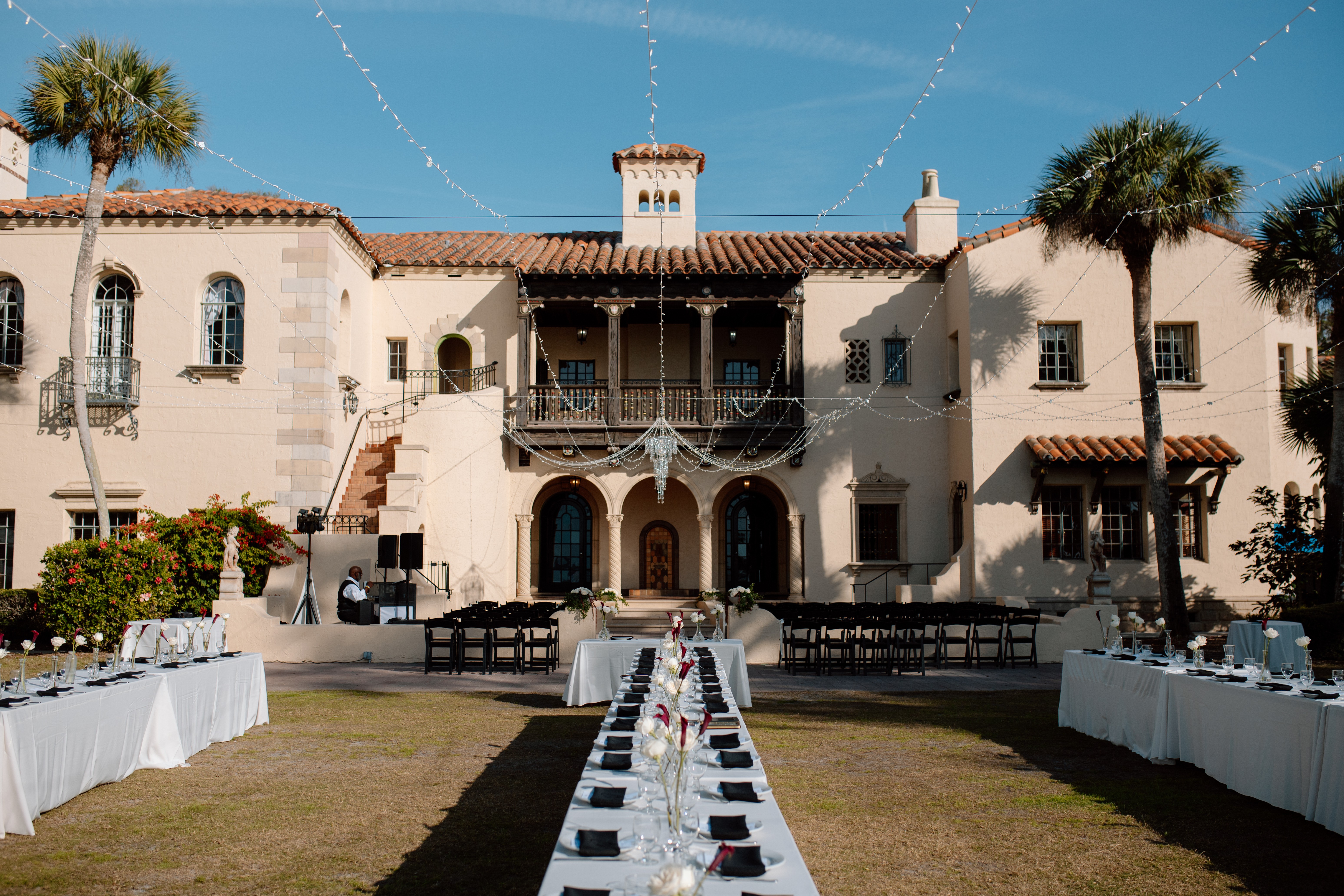 Large historic building with palm trees, string lights, and outdoor tables on lawn, viewed from front entrance.