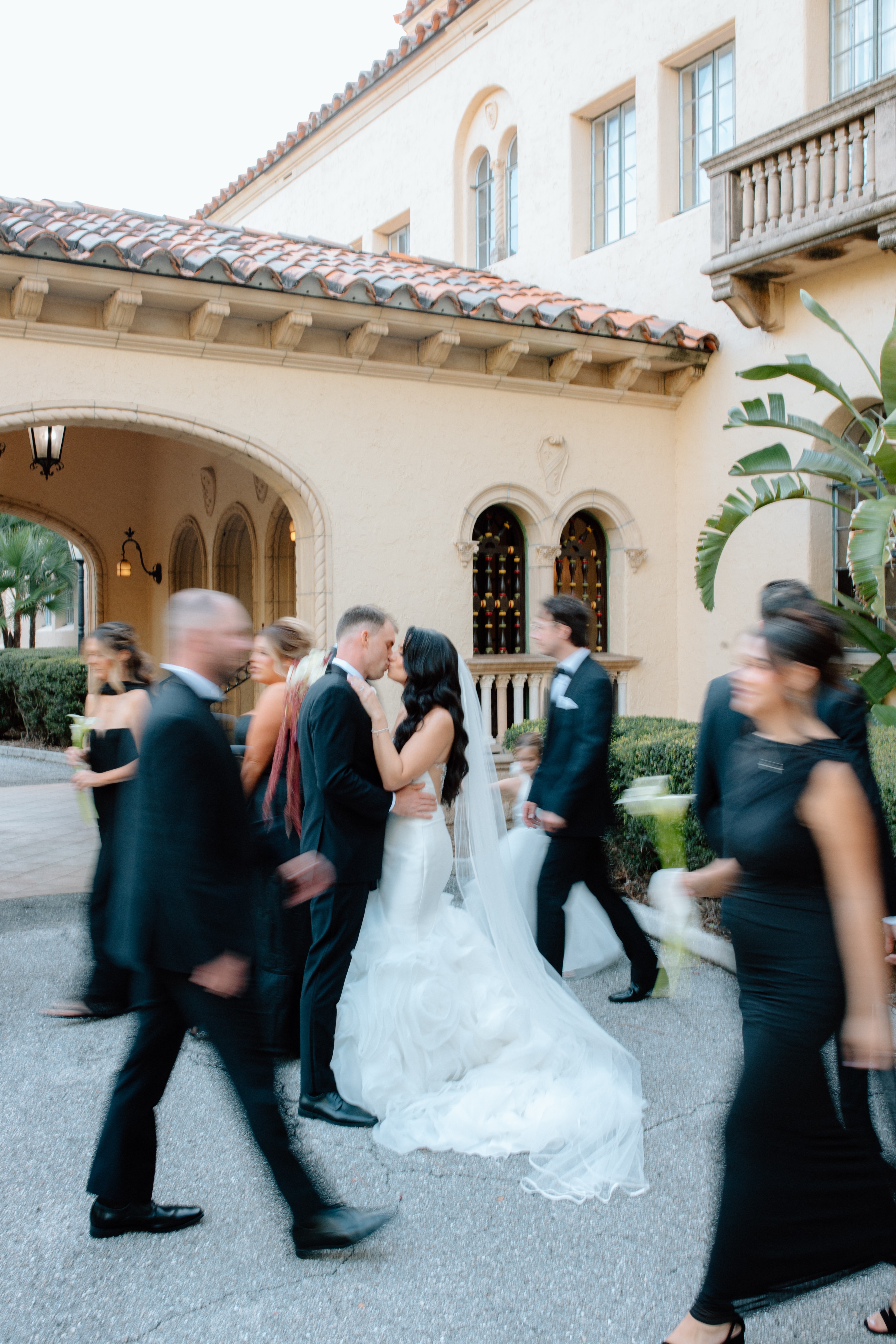 Couple in wedding attire kissing outside a building with people walking by, some blurred, in a courtyard setting.