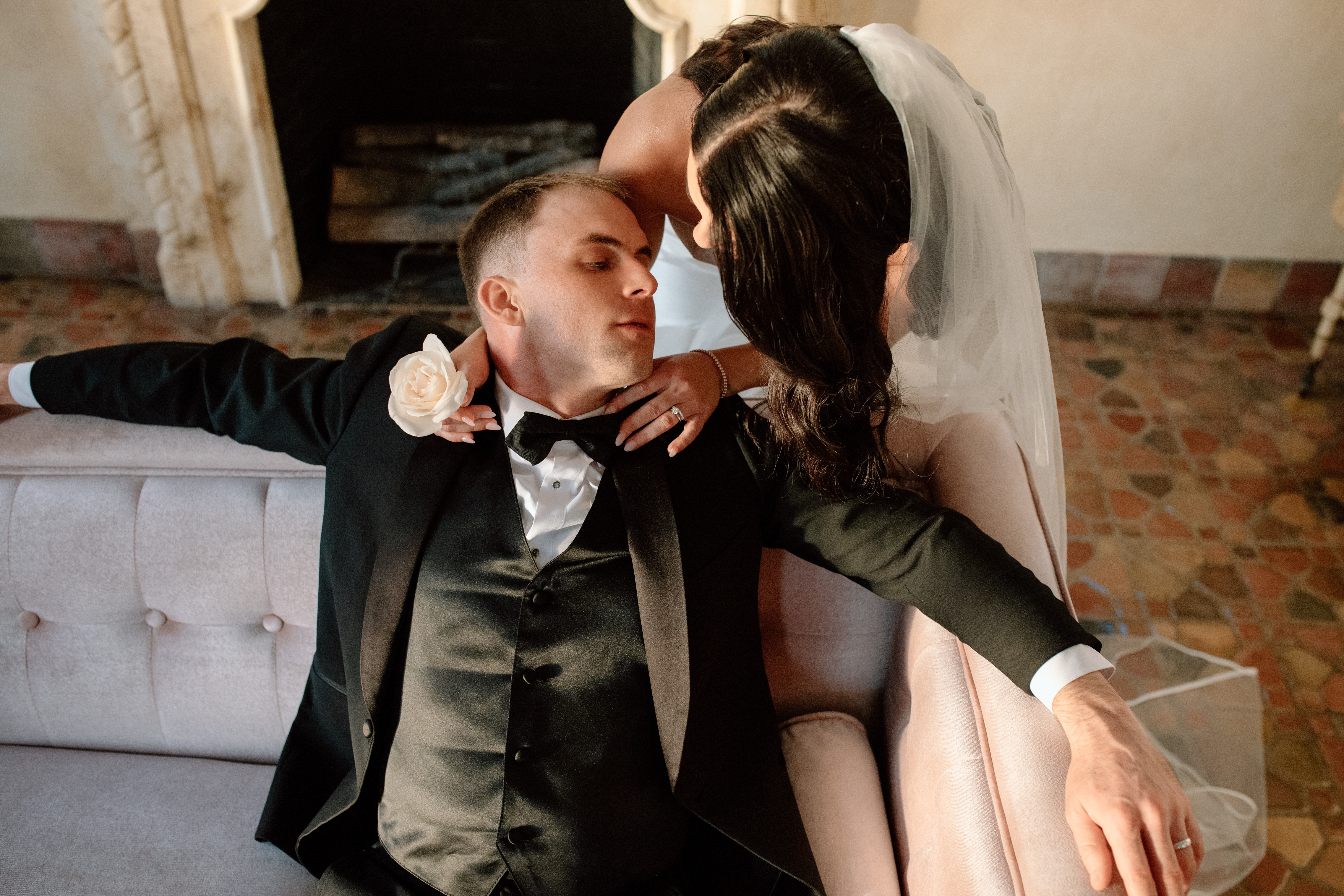 Bride and groom sitting close, bride leaning over groom, in a room with brick floor and fireplace, intimate moment.