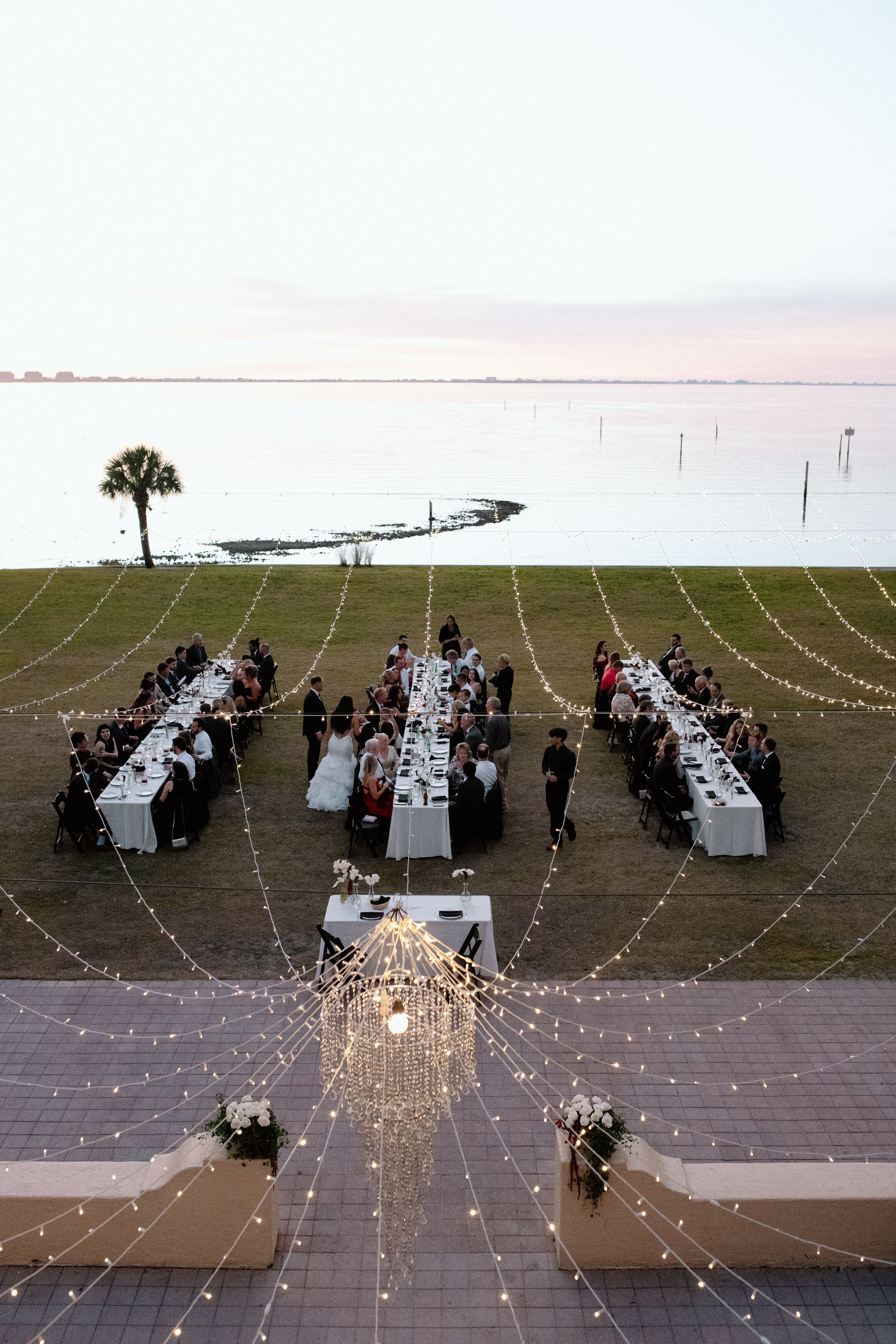 Outdoor event with long tables, people, and string lights near water, under a clear sky.