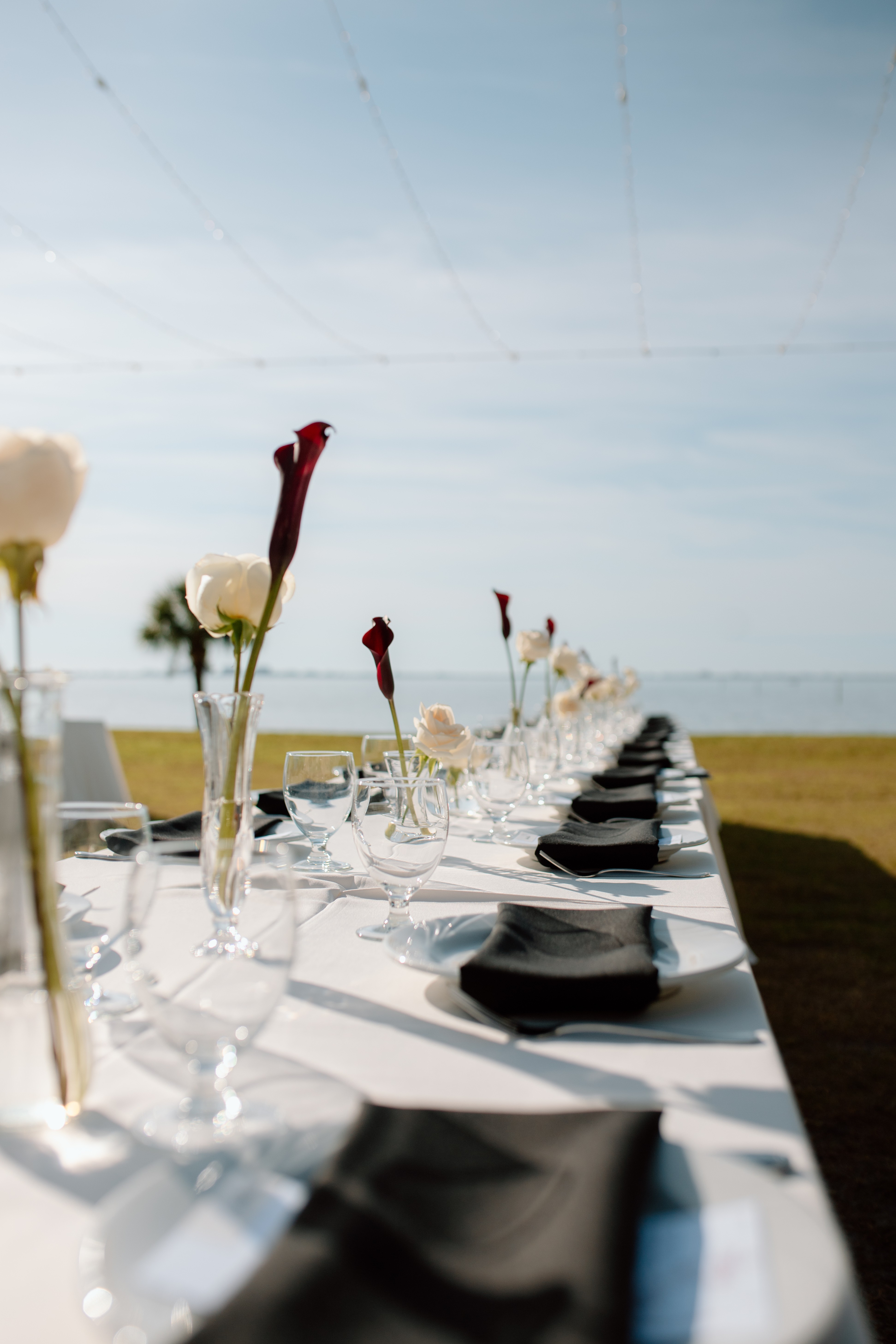 Long outdoor table set with black napkins, glassware, and tall vases with white and red flowers, overlooking a body of water.