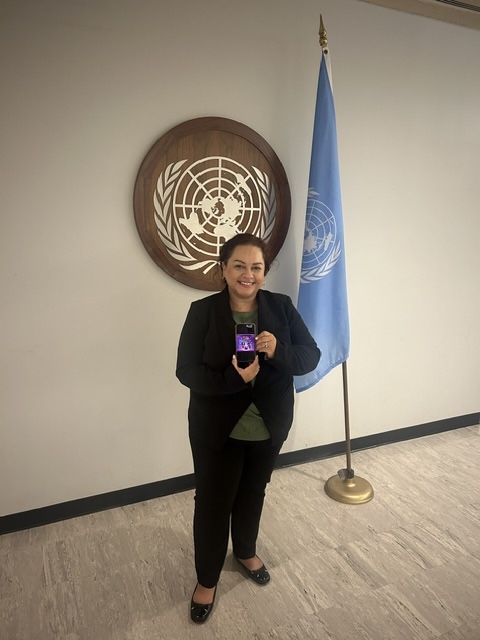 Woman in black blazer and pants taking a selfie in front of a United Nations emblem and flag, smiling.