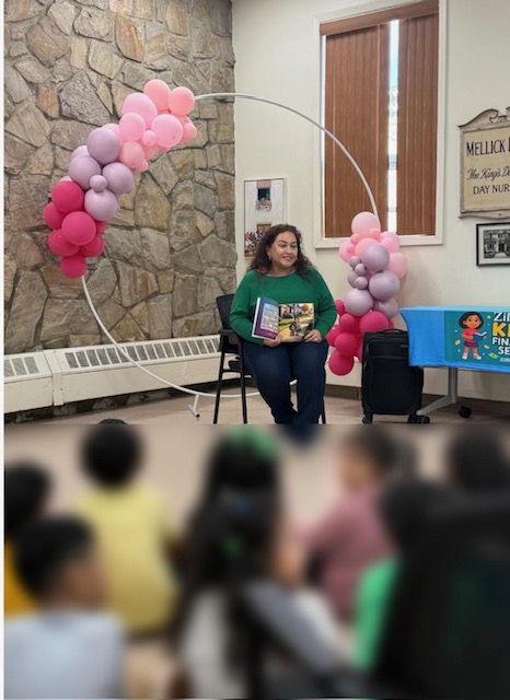 Woman sitting on a chair reading a book, decorated with pink and purple balloon arch, audience in foreground, indoor setting.