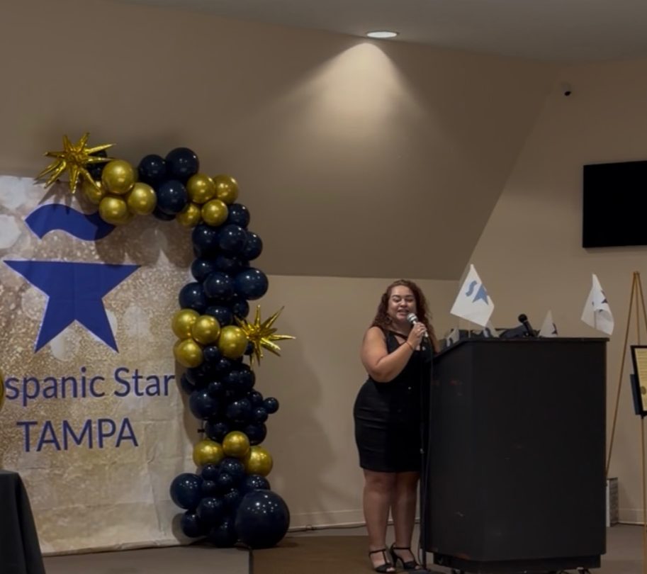 Woman speaking at a podium during an event with a decorated backdrop and balloon arch, in an indoor setting.