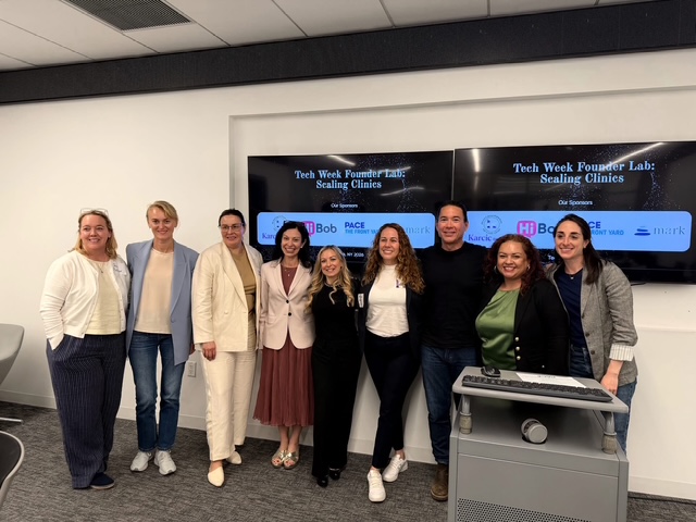 Group of nine women standing in front of a large screen displaying text about sealing clinics, smiling, in a conference room.